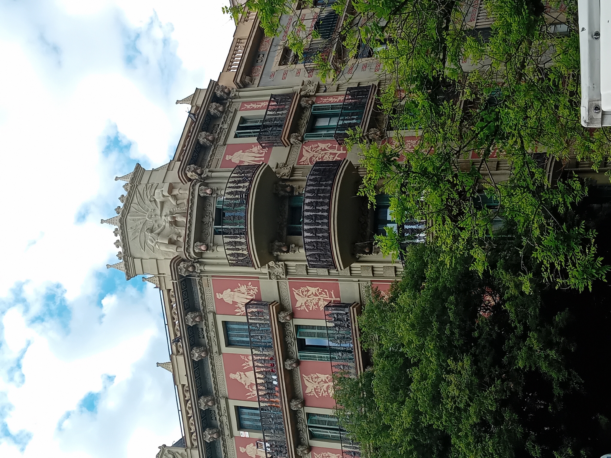 The image shows a building in Barcelona, Spain, viewed from below at an angle. The building is ornate, with decorative facades, sculpted details, and wrought-iron balconies. Its exterior is painted in tones of beige, brown, and red. The sky is visible in the upper left, with scattered clouds. A large green tree obscures part of the building on the right side of the frame. The building's architecture is in the modernist style, common to the city.