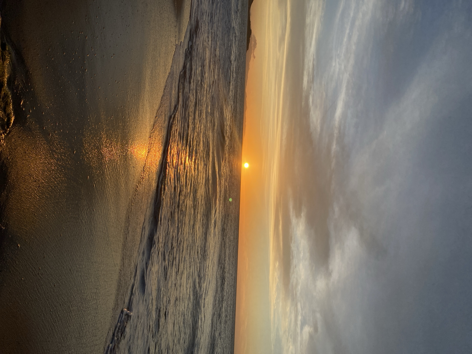 The image depicts a sunset at a beach. The scene is split vertically, with the beach and ocean on the left and the sky on the right. The sun is positioned at the center, casting a golden glow across the water and sand. The sky is filled with clouds, tinted with yellow and gray hues. The beach appears sandy, with gentle waves lapping the shore. No people or specific objects are clearly visible. The location is specified as Juan Griego, Venezuela, although this is not evident from the image itself.