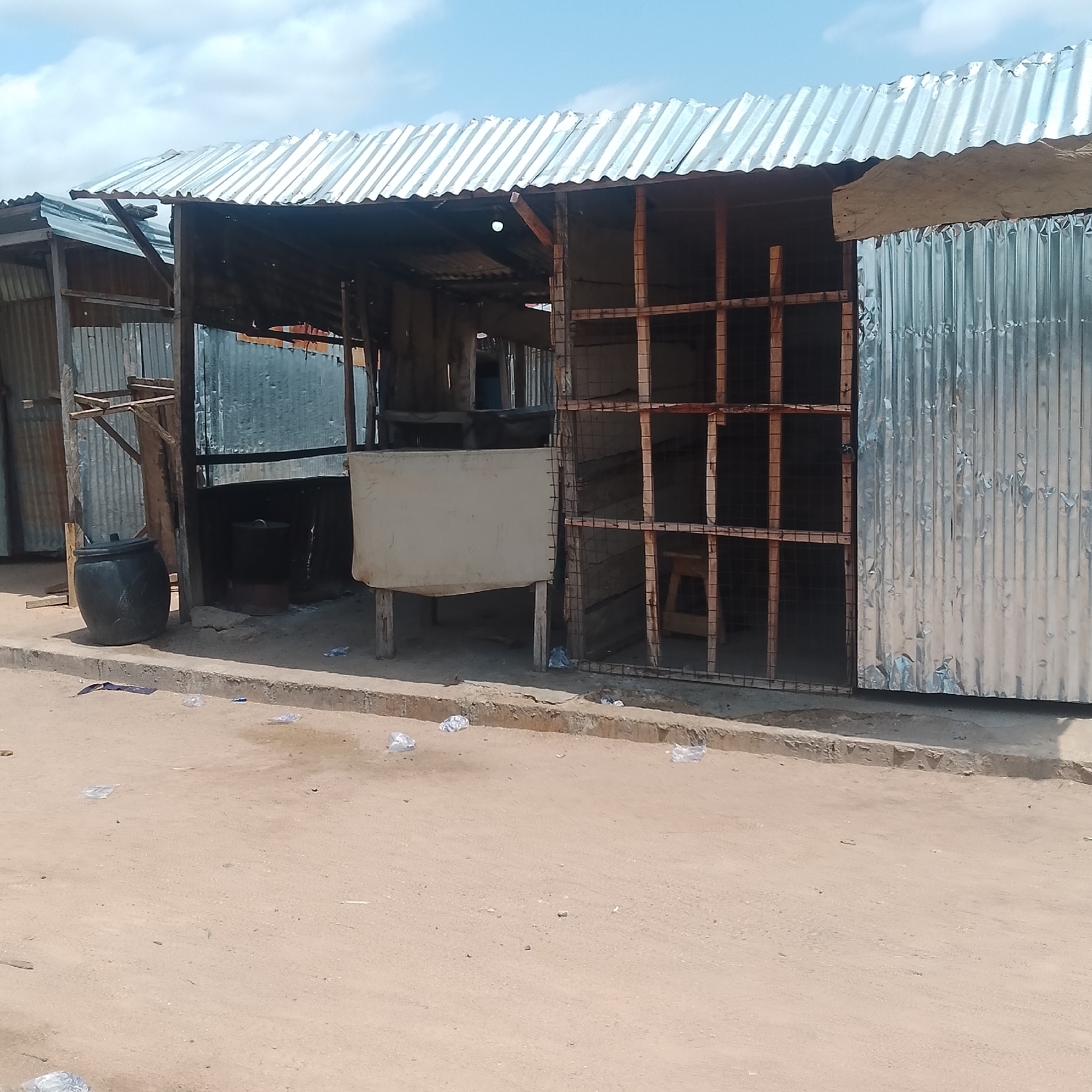 The image depicts a scene in Jalingo, Nigeria, featuring a makeshift structure constructed from corrugated metal and wood. The structure appears to be a market stall or shop, with an open front revealing an interior. Visible objects within the stall include a large black pot or container, a metal cage-like structure with wooden framing, and a light-colored fabric or tarp suspended on wooden poles. The exterior of the stall is made of corrugated metal sheeting, and the roof is also constructed from similar material. The ground in front of the stall is dirt and sand, with some scattered litter, including plastic bags and wrappers. A concrete curb or raised edge is present along the front of the stall. The overall impression is one of a simple, functional commercial space in an outdoor, possibly arid, environment.