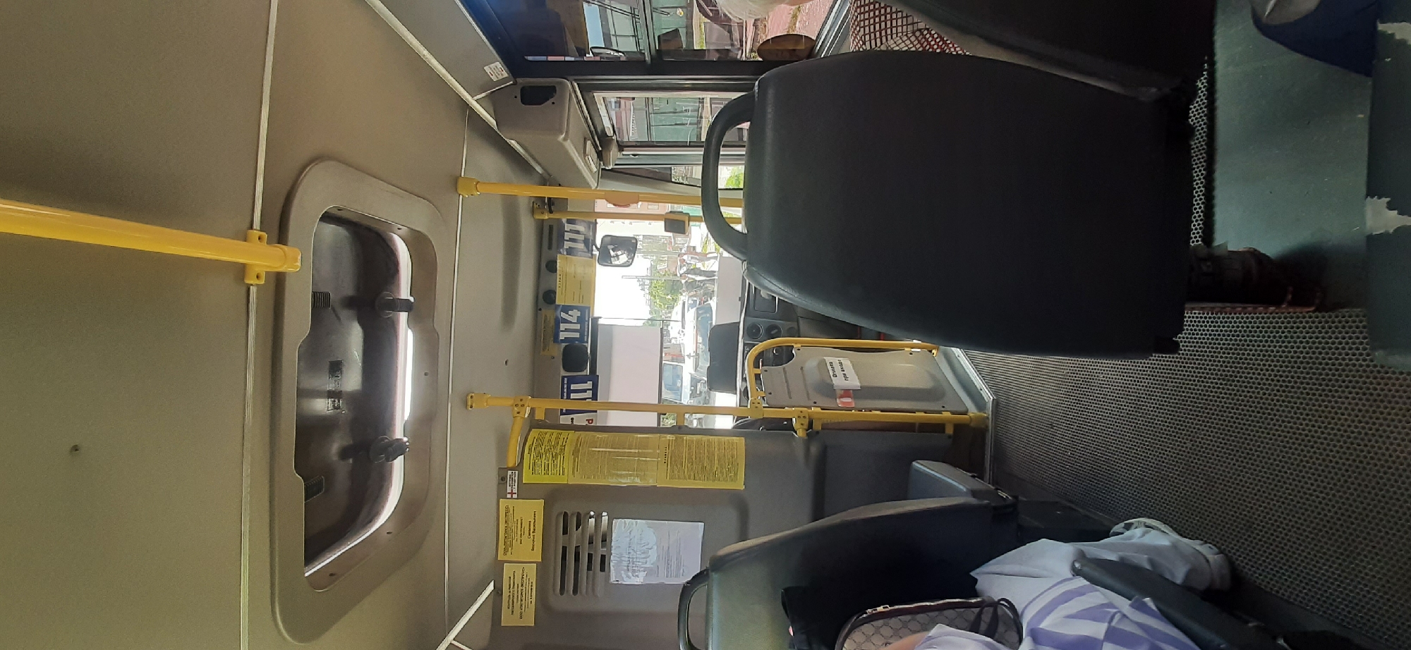 The image shows the interior of a bus, likely in Yessentukskaya, Russia, as indicated by the text on the signs within the bus which are in Russian and feature Cyrillic script. The bus is empty of passengers except for a partially visible person in the lower left corner, wearing a white and purple striped garment and a patterned bag. The bus has typical interior features: dark-colored seats, yellow handrails, a textured grey floor mat, and various signs mounted on the walls. One sign displays the number "114" in a blue rectangle, suggesting it is a route number. Another sign, red and white, reads "Оплата при входе" which translates to "Payment upon entry." The lighting suggests it is daytime, with sunlight entering through the windows. The overall impression is that of an ordinary public transport vehicle.