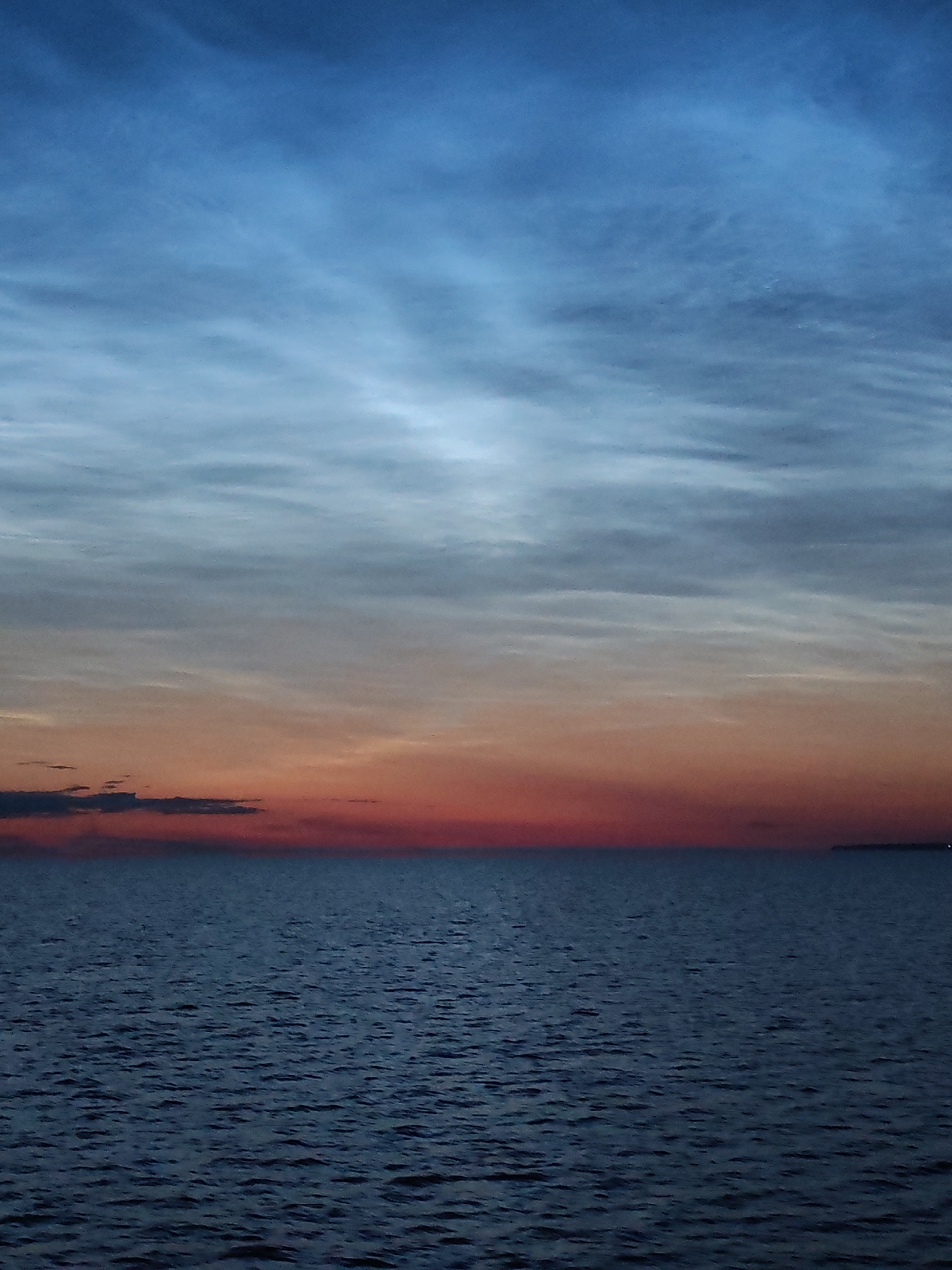 The image depicts a serene seascape at twilight, with a gradient of colors in the sky transitioning from deep blue at the top to fiery orange and red near the horizon. The water is a dark, rippling blue, reflecting the colors of the sky. The horizon is barely visible, with a faint suggestion of land or a distant object on the right. The scene is devoid of any human presence or man-made structures, creating a sense of vastness and tranquility. The location is identified as Ozernoe Ust'e, Russia, suggesting a coastal or lakeside setting. The overall atmosphere is one of peacefulness and natural beauty, characteristic of a sunset or sunrise over a body of water.