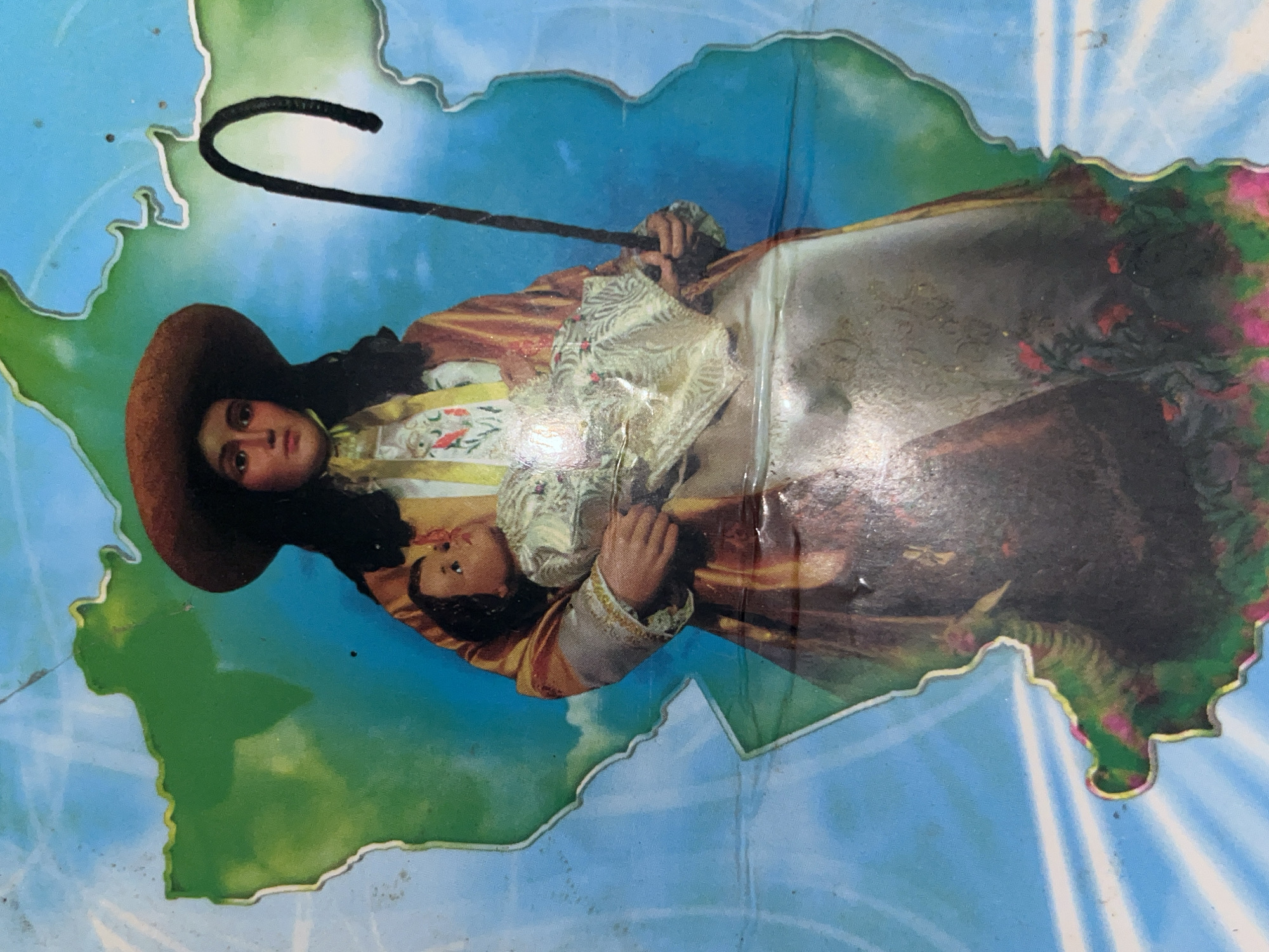 The image shows what appears to be a depiction of Divina Pastora, a religious figure, superimposed on an outline that somewhat resembles Venezuela. The figure, wearing a wide-brimmed hat and holding a staff, is depicted with a child. The background is blue with white radiating lines, and the outline of the figure is on a green field. Based on the Divina Pastora imagery, the location could be Tinaquillo, Venezuela, known for its devotion to the Divina Pastora.