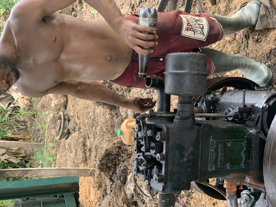 A shirtless man wearing red shorts and green rubber boots stands in front of a large, black, industrial-style engine. The man is holding a plastic bottle with some type of liquid in his right hand and an orange plastic container in his left hand. The engine is surrounded by dirt and what appears to be dried vegetation. The man appears to be working on the engine, possibly adding lubricant. The location is Egbeda, Nigeria, which is in the western part of the country. The image suggests the man is participating in a maintenance activity related to the engine.