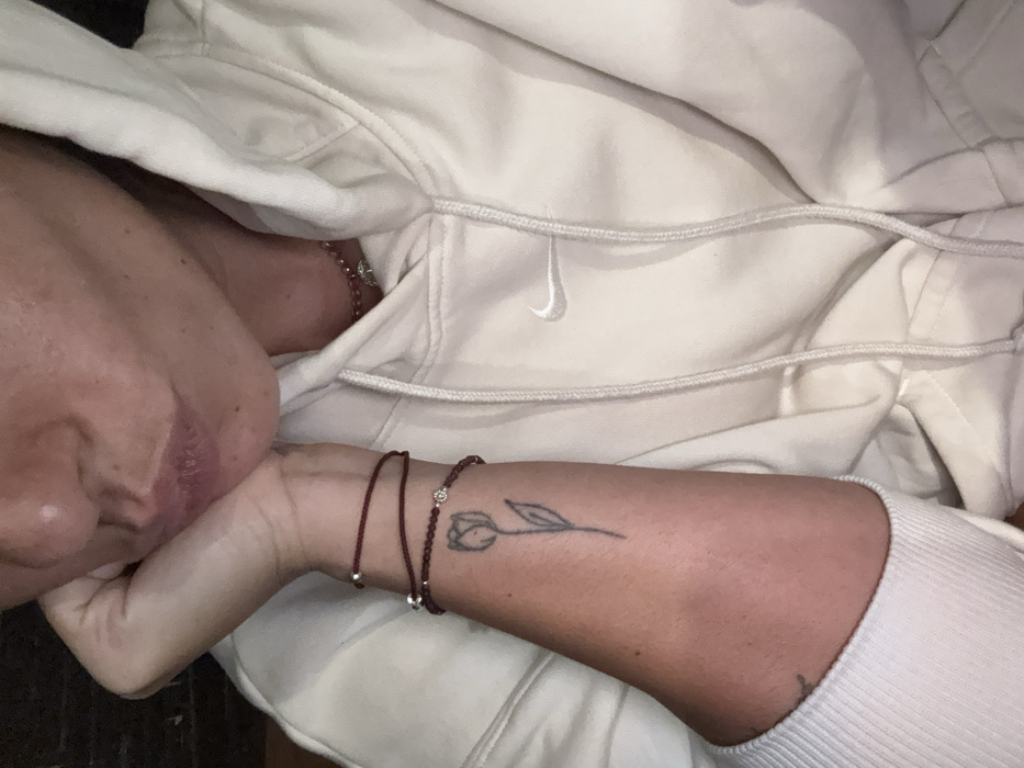 The image displays a close-up, cropped view of an individual, focusing on their lower face, right arm, and upper torso. The person's right hand is positioned beneath their chin. Their lips are slightly parted. The individual is wearing a light-colored, possibly off-white or cream-colored, hooded sweatshirt with a prominent white Nike swoosh logo stitched on the chest area. The sweatshirt features ribbed cuffs. On the right forearm, a simple black outline tattoo depicting a tulip flower with a stem is visible. Two dark red (maroon) bracelets adorn the right wrist: one is a thin, double-wrapped string or cord, and the other is a beaded bracelet with a single silver-colored, possibly faceted, bead or charm at its center. A thin, reddish beaded necklace with a small, circular, silver-colored pendant (resembling a tree of life design) is visible at the base of the neck. The background is dark and indistinct, offering no specific environmental details. The image does not provide visual cues to confirm a location in General Luna, Philippines.