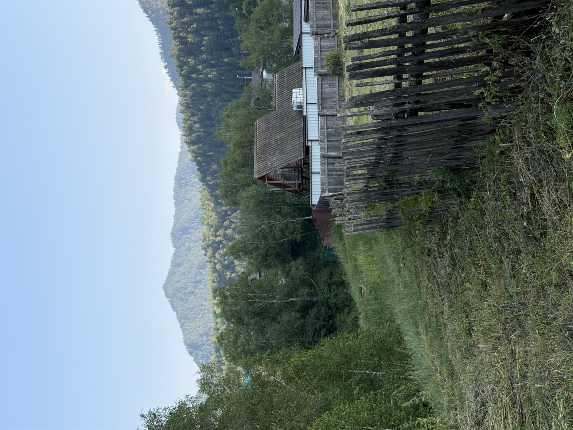 This is a vertical, eye-level, medium shot photograph taken in Anos, Russia. In the foreground, a rustic, weathered wooden fence made of vertical pickets runs diagonally from the lower right corner towards the center left of the frame. Beyond the fence, a grassy area with some recently cut vegetation is visible. In the mid-ground, a large wooden house with a corrugated roof stands behind a light-colored metal fence. The house has a balcony and appears to be elevated. To the left of the house, a dense forest of deciduous and coniferous trees extends into the background. Further in the distance, rolling, forested hills rise up, meeting a clear, pale blue sky. The overall scene depicts a rural residential area nestled within a mountainous, forested landscape.