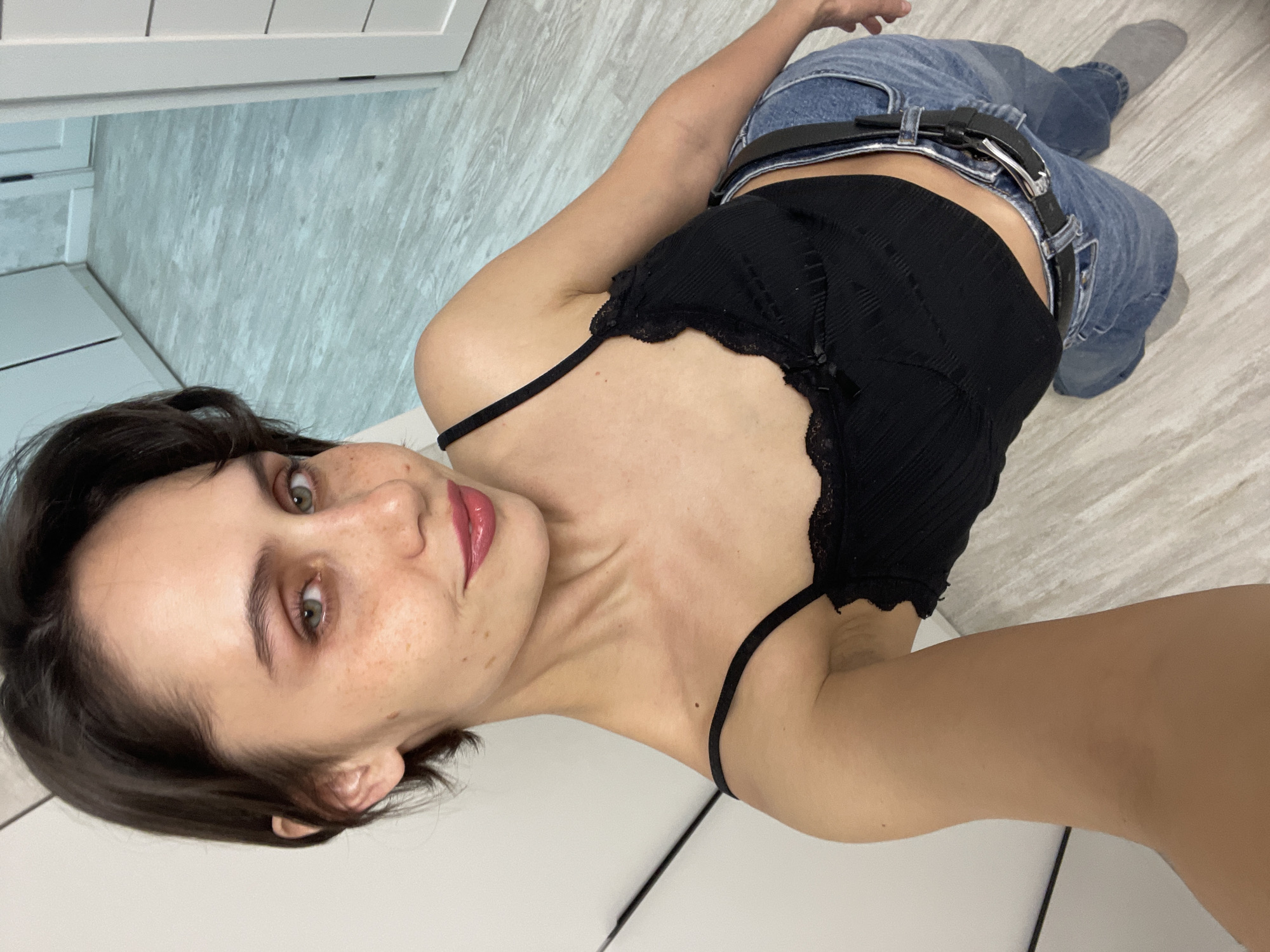 A young woman is depicted lying on a light-colored tiled floor, looking directly at the camera with a subtle smirk. Her body is oriented vertically in the rotated image. She has short, dark brown hair, light-colored eyes (appearing greenish-hazel), and freckles across her face. Her lips are adorned with glossy pink lipstick.
She is wearing a black, ribbed or knitted sleeveless crop top with delicate lace trim and thin spaghetti straps. Her lower body is covered by blue denim jeans, cinched with a black belt featuring a prominent silver buckle. One foot is visible, wearing a grey sock.
The immediate setting is an indoor space with a clean, light-colored tiled floor. In the background, there are white cabinets or furniture and a different section of flooring, which is light grey with a textured pattern. The lighting suggests it is daytime. The woman appears to be posing for a photograph. The scene is situated in Izhevsk, Russia. No visible text is present in the media.
