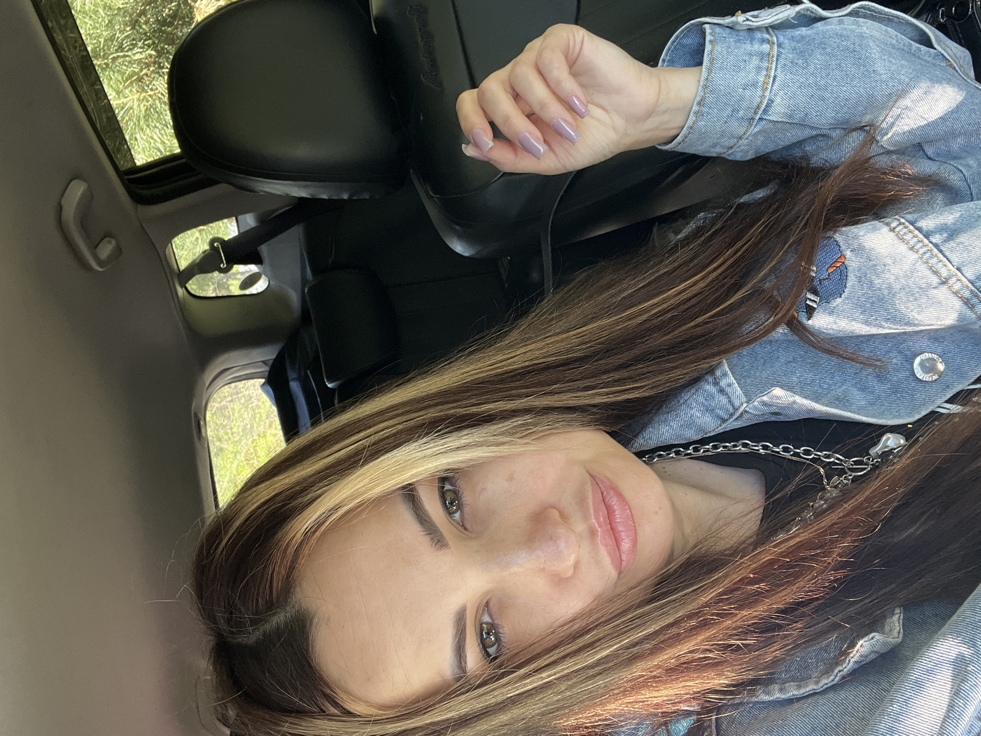 The image shows a woman in a car. She has long, brown hair with blonde highlights and is wearing a denim jacket and a silver chain necklace. Her nails are painted a light purple color. The interior of the car is visible, including the black leather seats and the sunroof. The background shows trees and greenery outside the car window. The exact location in the city of Unknown, Unknown is not discernible from the image.