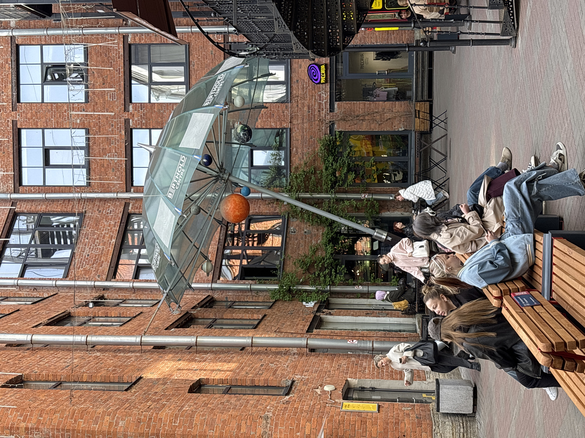 The photograph depicts a group of people reclining on benches in what appears to be a courtyard in Saint Petersburg, Russia. The area is enclosed by a brick building with multi-paned windows and a metal umbrella-like structure adorned with spheres of varying sizes. Overhead, ornate metalwork adds to the architectural detail.
Several individuals are lying on the wooden benches, many with their legs extended. The seated people appear relaxed and engaged in conversation. The area is paved with red and grey bricks, and there's evidence of commercial activity, with a storefront visible in the background. The scene suggests a leisure or gathering spot within a city setting.
