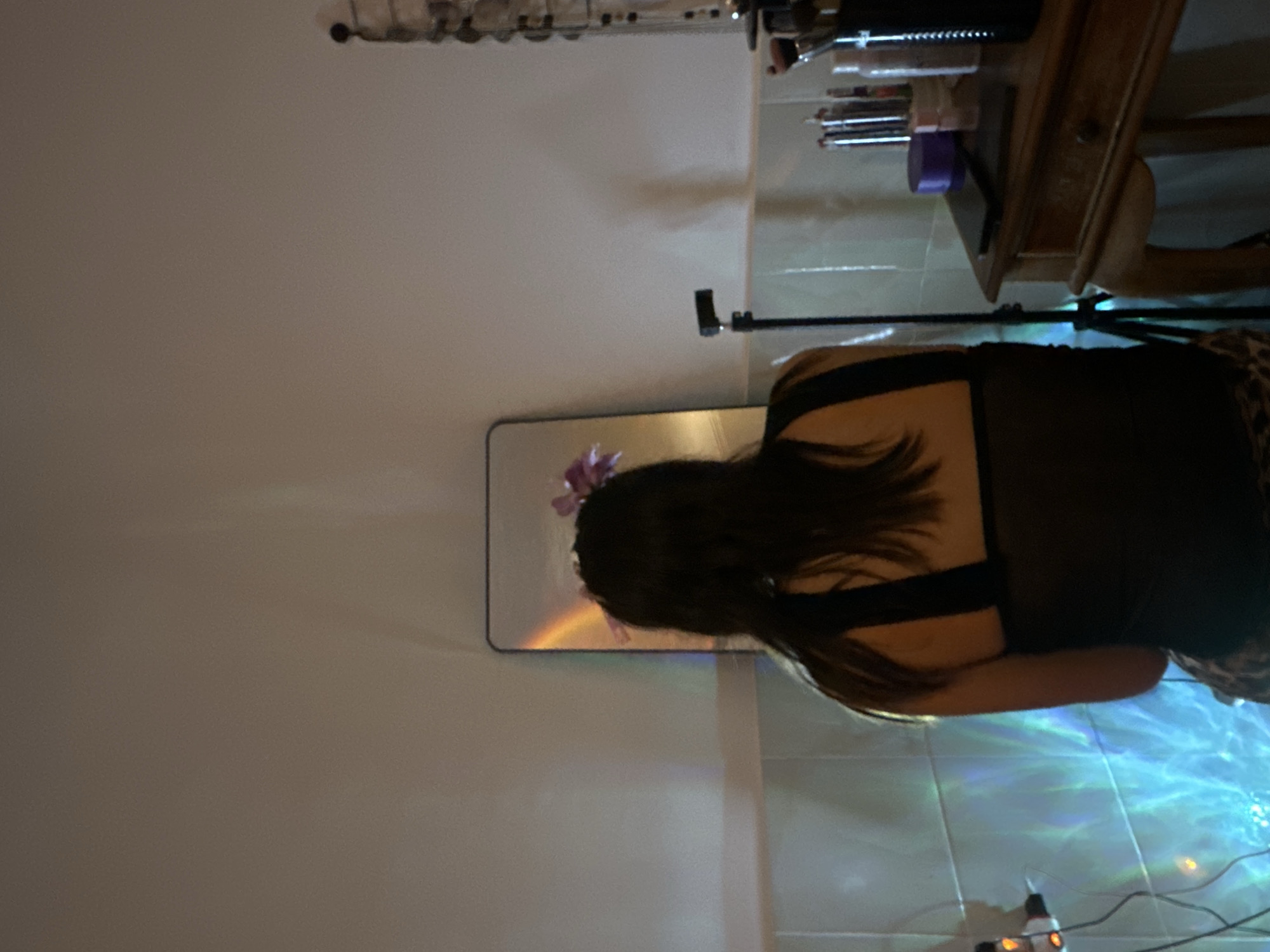 The image displays an indoor scene featuring a person seen from behind, sitting or leaning while facing a rectangular mirror. The individual has long, dark hair and is wearing a dark top. In the reflection within the mirror, a distinctive purple flower or decorative accessory is visible on the person's head.
The setting appears to be a room characterized by light-colored walls and tiled flooring. Towards the upper right side of the frame, a wooden table or shelf is visible, adorned with various items such as makeup brushes and bottles. A black stand, likely a tripod or a light stand, is positioned in the mid-ground.
A notable detail is the floor directly in front of the person, which is illuminated by dynamic blue and green light patterns. These patterns evoke the appearance of moving water reflections or the effect of a light projector, contributing to an ethereal and vibrant atmosphere. A small white plug and some electrical wires are also visible on the floor.
The activity suggested is personal grooming, such as applying makeup or styling hair, possibly in preparation for an event or a photo opportunity. The overall ambient lighting appears dim, which accentuates the colorful projected lights. There is no visible text, and no specific emotions are discernible from the person's back.