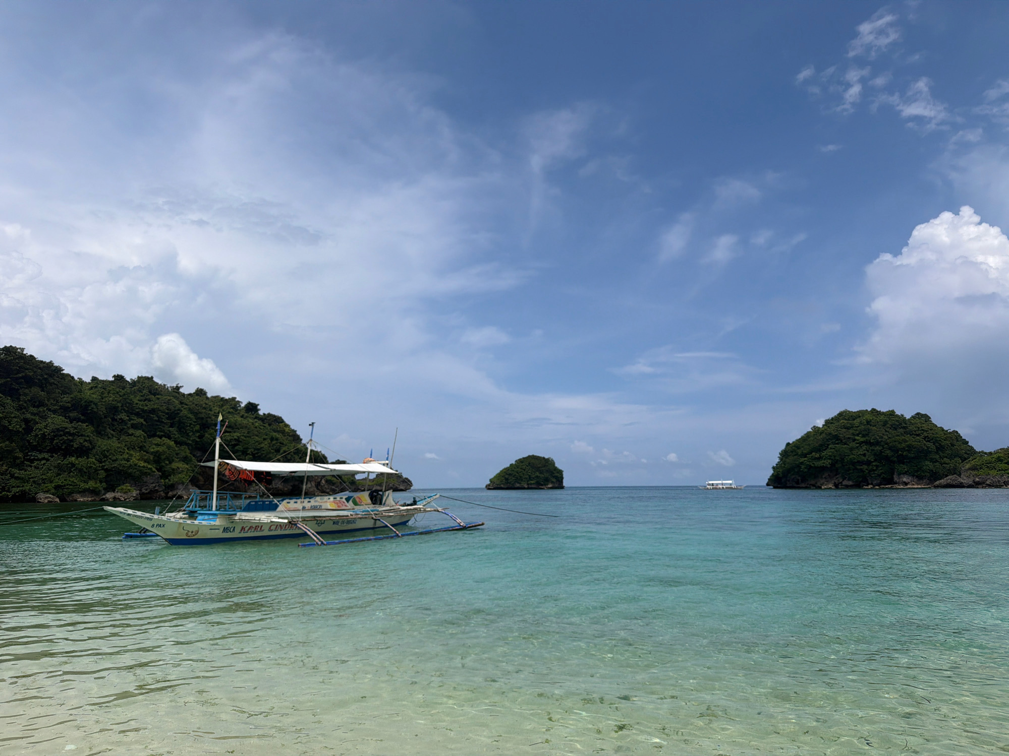 The image depicts a tropical seascape with a clear turquoise ocean and a bright blue sky dotted with white clouds. In the foreground, the shallow, sandy bottom of the ocean is visible through the crystal-clear water. A traditional Filipino outrigger boat, named "MSCA KARL CINDERELLA" and identified to carry "8 PAX", is anchored in the water, its blue and white hull contrasted by colorful decals. The boat has a white canopy providing shade. To the left, a lush, green, tree-covered island rises from the water, suggesting a coastal or island location. In the distance, two smaller, verdant islets are visible, and further out, another white boat is seen on the horizon. The overall scene conveys a sense of tranquility and natural beauty, consistent with popular tourist destinations in the Philippines. This scene is representative of locations in Malay, Philippines, a municipality in the province of Aklan, known for its beautiful beaches and islands.