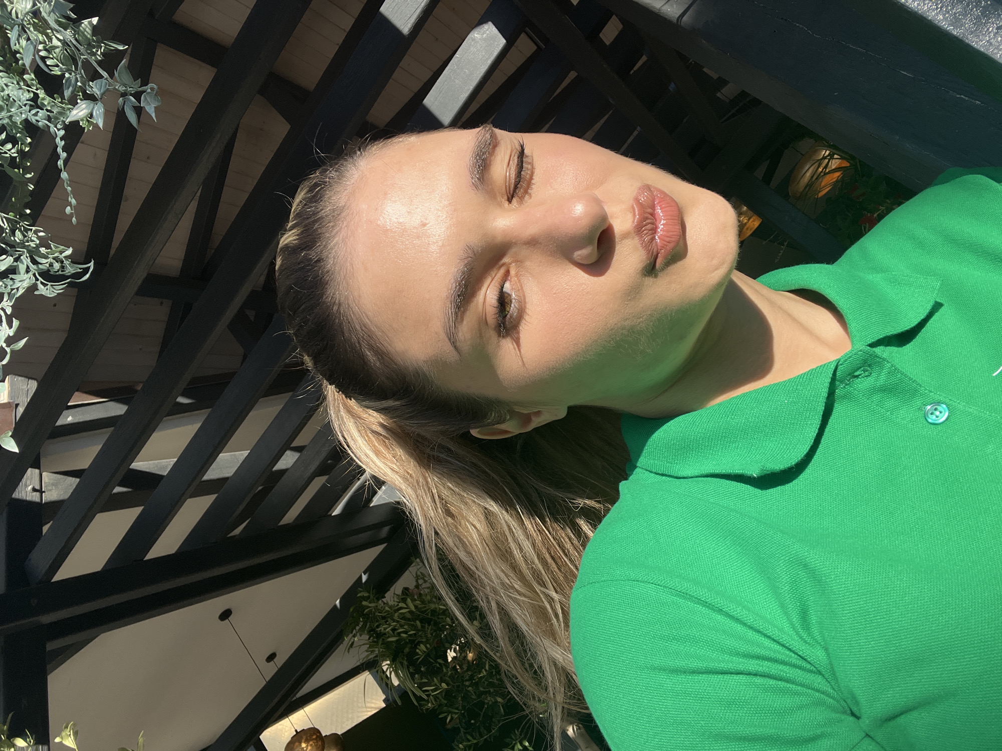 The image is a close-up, low-angle selfie of a young woman with her eyes closed and lips pursed. She has long, blonde hair tied back in a ponytail, and she is wearing a bright green polo shirt. The lighting suggests it is daytime, with sunlight casting shadows from a dark, slatted overhead structure, likely a pergola or canopy. Green foliage is visible to the left and bottom right of the frame, and behind her, the background is blurred, showing hints of plants and possibly outdoor lighting fixtures. The woman's skin appears dewy, and her lips have a glossy finish. The overall impression is of a casual, sunny outdoor setting, possibly in a garden or patio area. There is no visible text in the image.