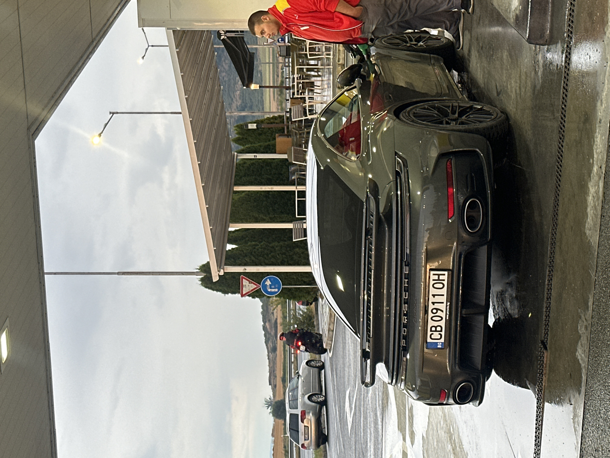 At a gas station in Drazhevo, Bulgaria, under a covered canopy, a dark grey Porsche 911 Turbo S is parked at a fuel pump. A man wearing a red and yellow jacket and grey pants stands by the car, appearing to be fueling it. The ground is wet and reflective, suggesting recent rain. In the background, other vehicles are visible, including a silver station wagon and a motorcycle with a rider. The environment appears to be an open area with trees and some distant hills under a cloudy sky. The license plate of the Porsche reads "CB 0911 OH".
