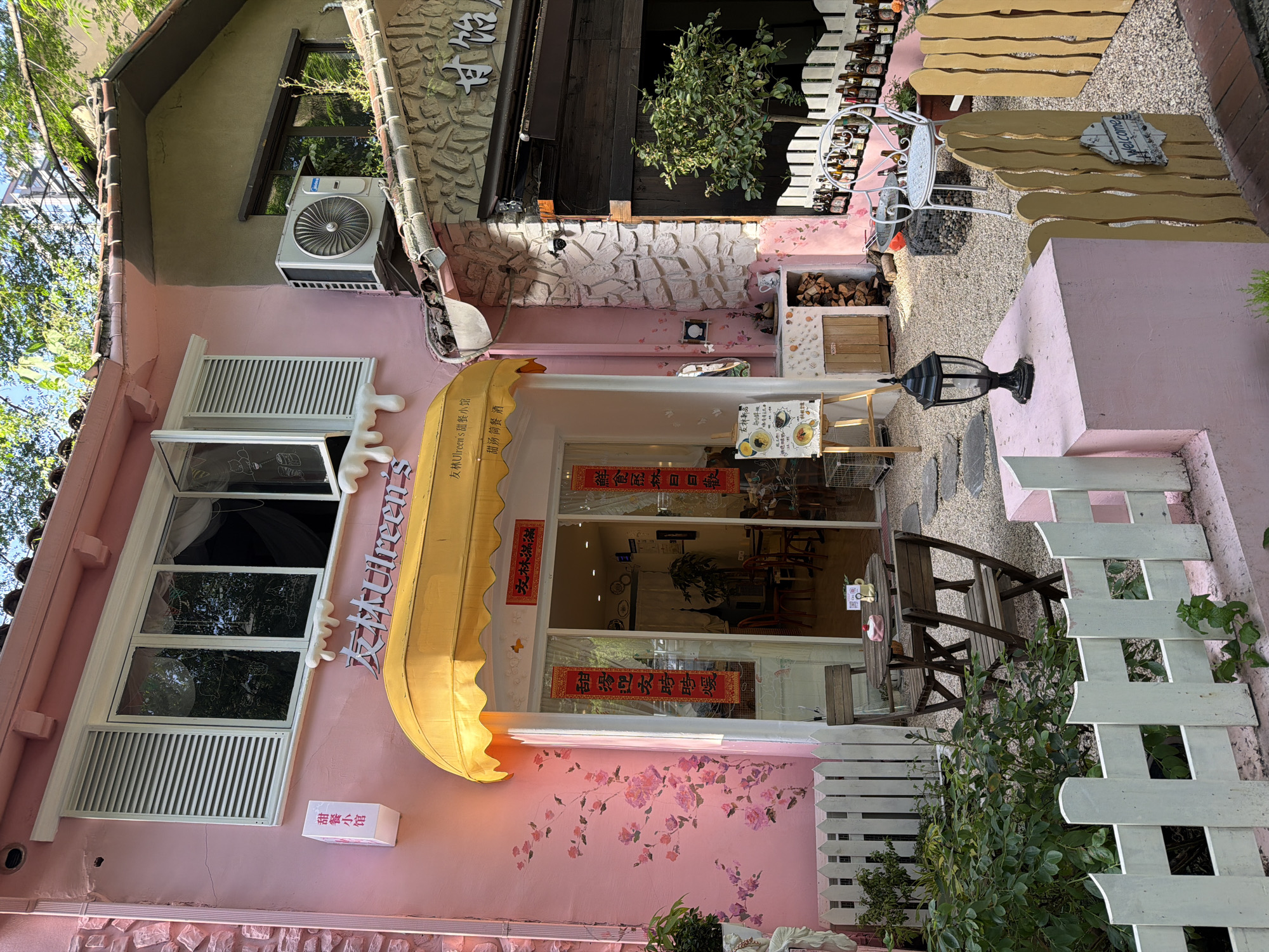 The image shows the exterior of a business, likely a cafe or restaurant, in Liangjiang, China. The building is painted pink and decorated with floral designs. The entrance is marked by a yellow awning with Chinese writing, partially obscuring the sign which reads "友林Ulreen's 甜餐小馆", indicating the business name is "Youlin Ulreen's Sweet Meal House". The entrance is flanked by signs with Chinese characters, and there are windows to the side with white frames and partially opened shutters. An air conditioning unit is mounted on the wall above the window. Outside, there are tables and chairs arranged on a gravel surface, as well as decorative elements like a small white fence and a welcome sign.
