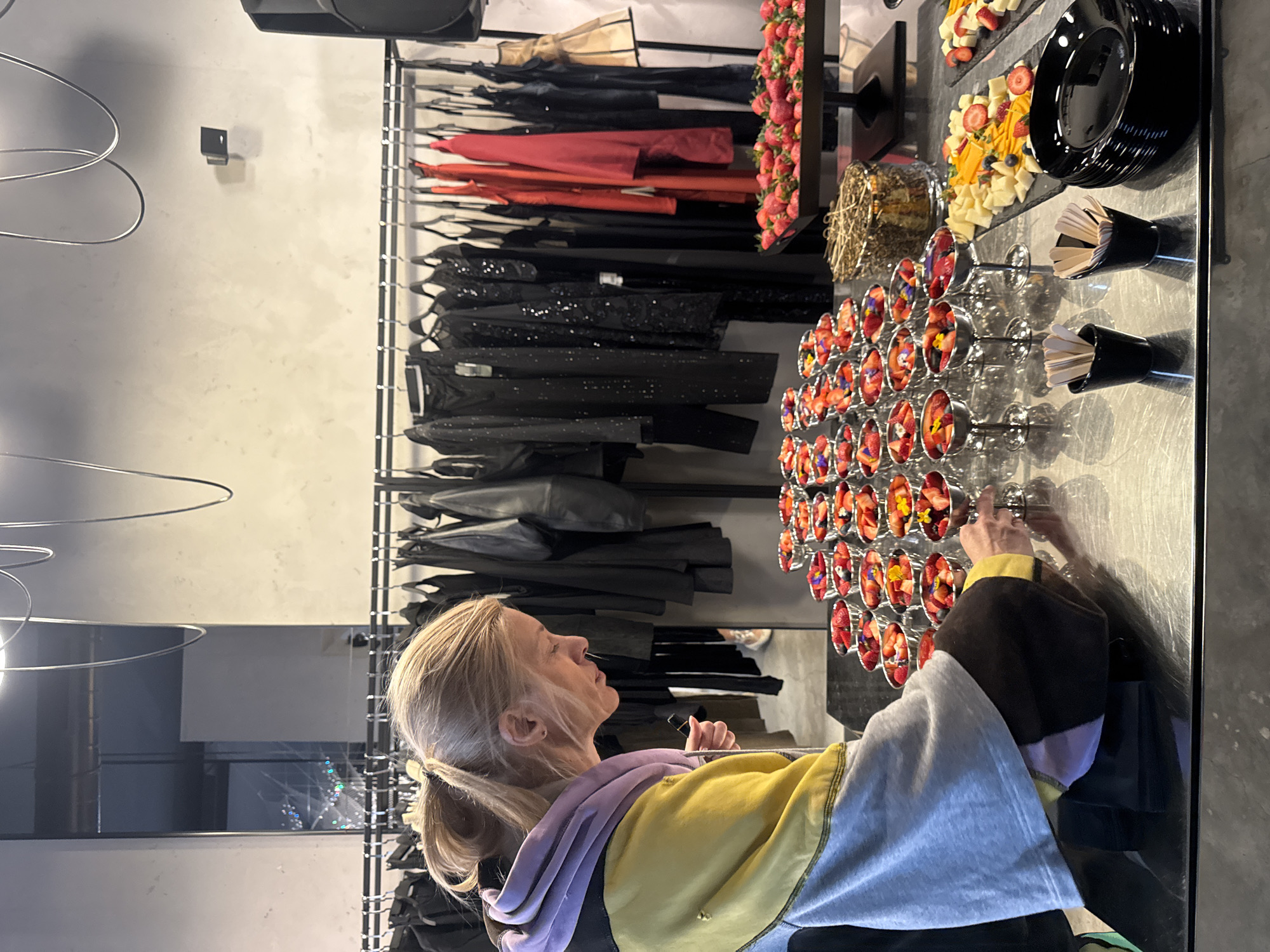 The image captures an indoor retail or event space located in Sofia, Bulgaria, featuring a minimalist industrial design. A blonde female, with her hair styled in a ponytail, is observed in profile on the left side of the frame. She wears a multi-panel hooded sweatshirt in shades of purple, yellow, gray, and dark brown/black. The woman is leaning forward, directing her gaze and right hand towards an elaborate food display arranged on a dark, reflective metallic counter.
The central feature of the food display is a tiered arrangement of approximately 30-40 individual silver-colored goblets, each containing mixed berries (primarily strawberries and blueberries) and garnished with small edible flowers. To the right, a black rectangular tray is piled high with fresh strawberries. Adjacent to this are multiple black slate-like boards presenting cubed cheeses, sliced fruit such as mango, strawberries, and blueberries. A round dessert, possibly a cake, adorned with gold-colored wire decoration, is also visible. A stack of black oval plates and two small black containers holding wooden disposable utensils complete the counter setup.
The background showcases multiple clothing racks against a light gray wall, displaying a collection of mostly dark-colored apparel including dresses, jackets, and tops, interspersed with a few vivid red garments. A large black audio speaker is positioned on the far right. On the left, a tall, narrow mirror reflects a blurred exterior nighttime street scene, showing vehicle lights and urban structures, suggesting a window view from the venue.