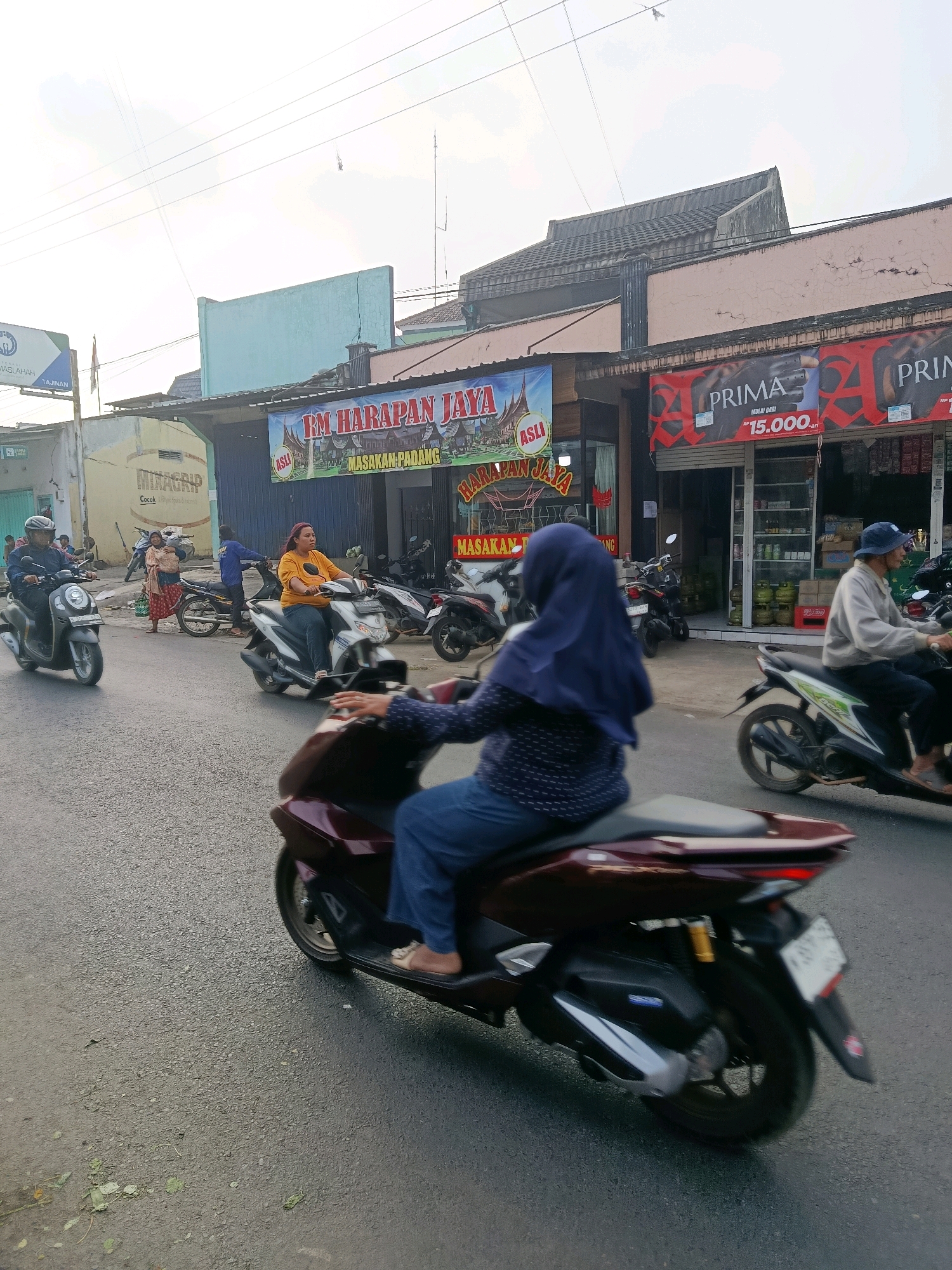 The image depicts a street scene in Tajinan, Indonesia, on a bright, possibly overcast day. The foreground features a maroon scooter with a person wearing blue jeans and flip-flops riding it. The scooter's taillight is illuminated. To the right, another scooter with a rider wearing a blue hijab and a patterned shirt is visible, also in motion. In the midground, a woman in an orange shirt and blue jeans rides a white scooter. Behind her, a man in a blue jacket stands near a motorcycle. Further back, another scooter carries a person in a blue hijab. The background shows a row of buildings with storefronts. One prominent banner reads "RM HARAPAN JAYA MASAKAN PADANG," suggesting a Padang cuisine restaurant. Another banner displays "PRIMA" with a price of "15.000." A smaller sign for "MIXAGRIP" is also visible. Several other motorcycles are parked or in motion. The street surface is asphalt with some scattered leaves and debris. Overhead, power lines crisscross the sky, and a cell tower is visible in the distance. The overall impression is of a bustling local street with moderate traffic.