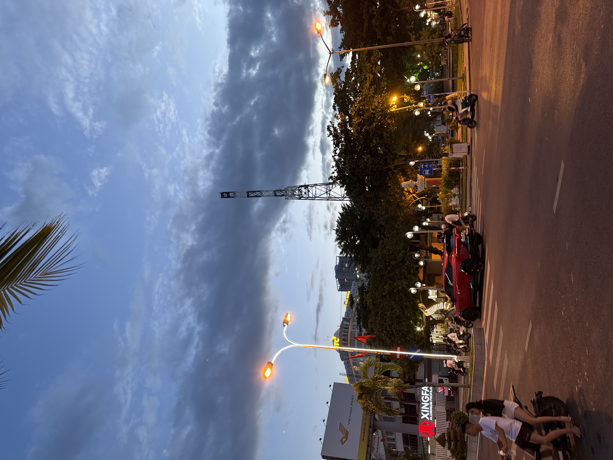 In Nha Trang, Vietnam, the scene depicts a street intersection during twilight. A tall, lattice radio tower stands prominently against a dramatic sky filled with dark, heavy clouds. In the foreground, streetlights cast an orange glow on the asphalt road. A red SUV is stopped at the intersection, and several motorcycles navigate the traffic. Two young people are riding on a motorcycle, with the passenger looking back. Buildings and trees line the street, some illuminated by exterior lights. A billboard for "Vietnam Post" is visible on the left side of the image. The overall atmosphere is that of a bustling city transitioning from day to night.