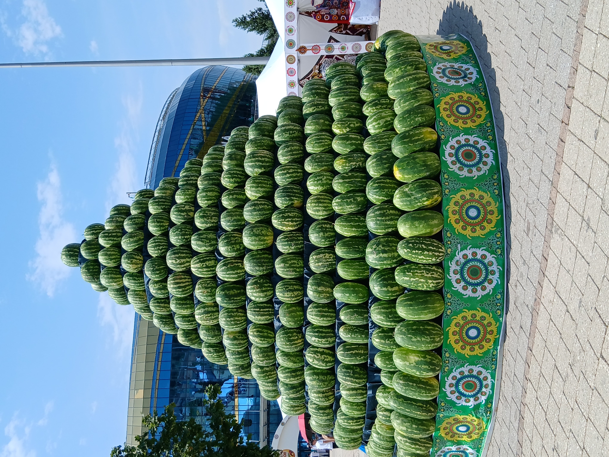 The image displays a large, pyramid-shaped arrangement of watermelons outdoors. The watermelons are stacked in many rows, forming a triangular structure that tapers towards the top. The arrangement is decorated with a vibrant green cloth featuring colorful, circular patterns. The scene appears to be set at a market or festival, possibly during a summer day, as indicated by the bright sunlight casting shadows on the paved ground. In the background, a modern building with a glass facade is visible, and beyond that, a clear blue sky with a few wispy clouds. The overall impression is one of abundance and celebration, with the neatly arranged watermelons suggesting a harvest festival or a display of local produce. There are no visible people prominently in the frame, though there are hints of tents and structures that might suggest a public gathering. The time of day seems to be daytime, likely mid-morning or afternoon, given the bright sunlight. The weather appears to be clear and sunny.