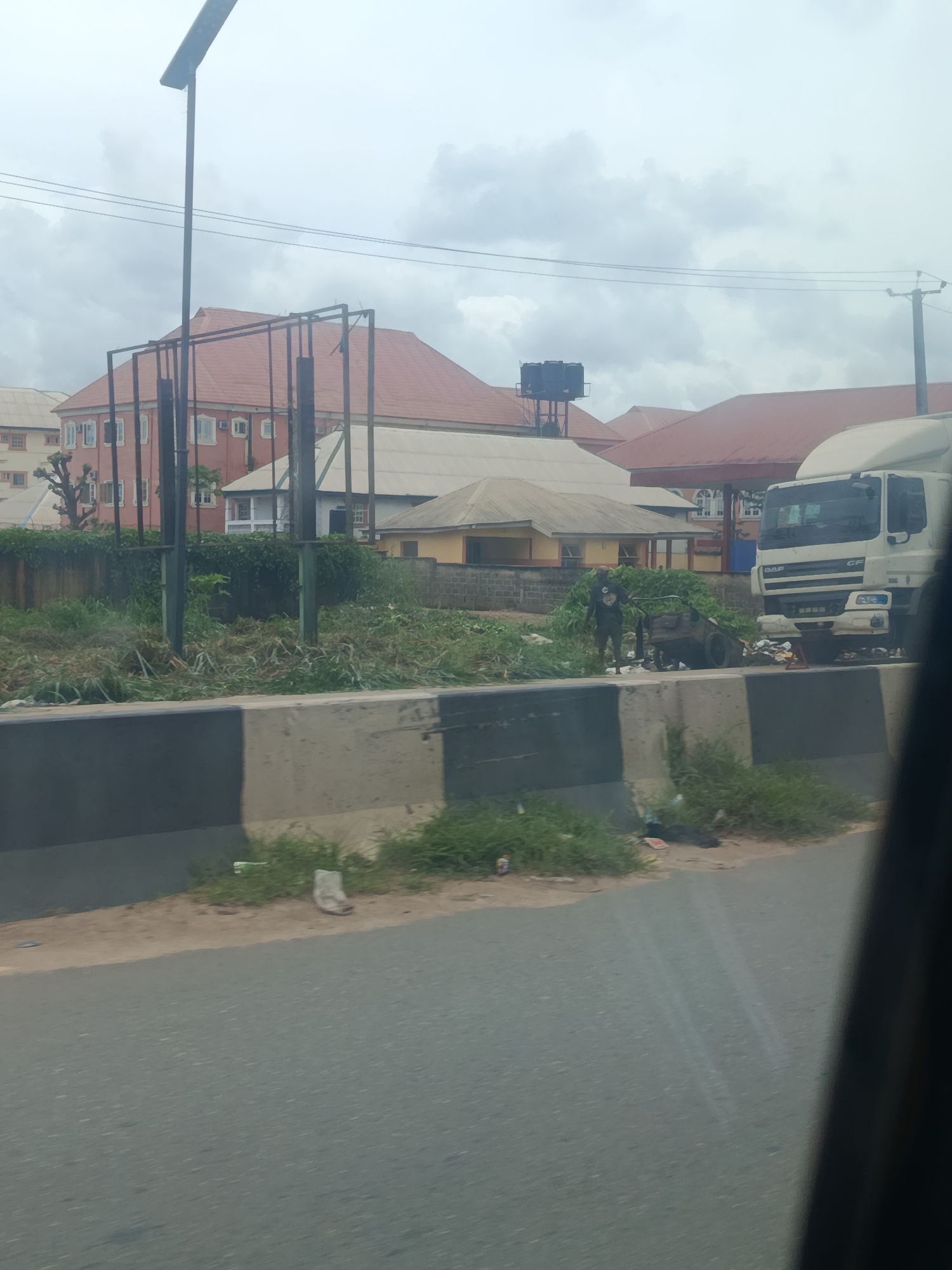 The image depicts a scene in Ogbaku, Nigeria, with a focus on an area adjacent to a road. In the foreground, a concrete barrier with black and white stripes runs horizontally. Beyond the barrier, a patch of overgrown vegetation and debris is visible. A man in dark clothing stands near a partially visible wheelbarrow and scattered trash. To the right, a white truck with "DAF" branding is parked, also surrounded by debris. In the background, several buildings with red-tiled roofs are visible, along with a tall street light and power lines. The sky is overcast with clouds. The overall impression is one of an urban or semi-urban environment with evidence of litter and disarray in the depicted area.