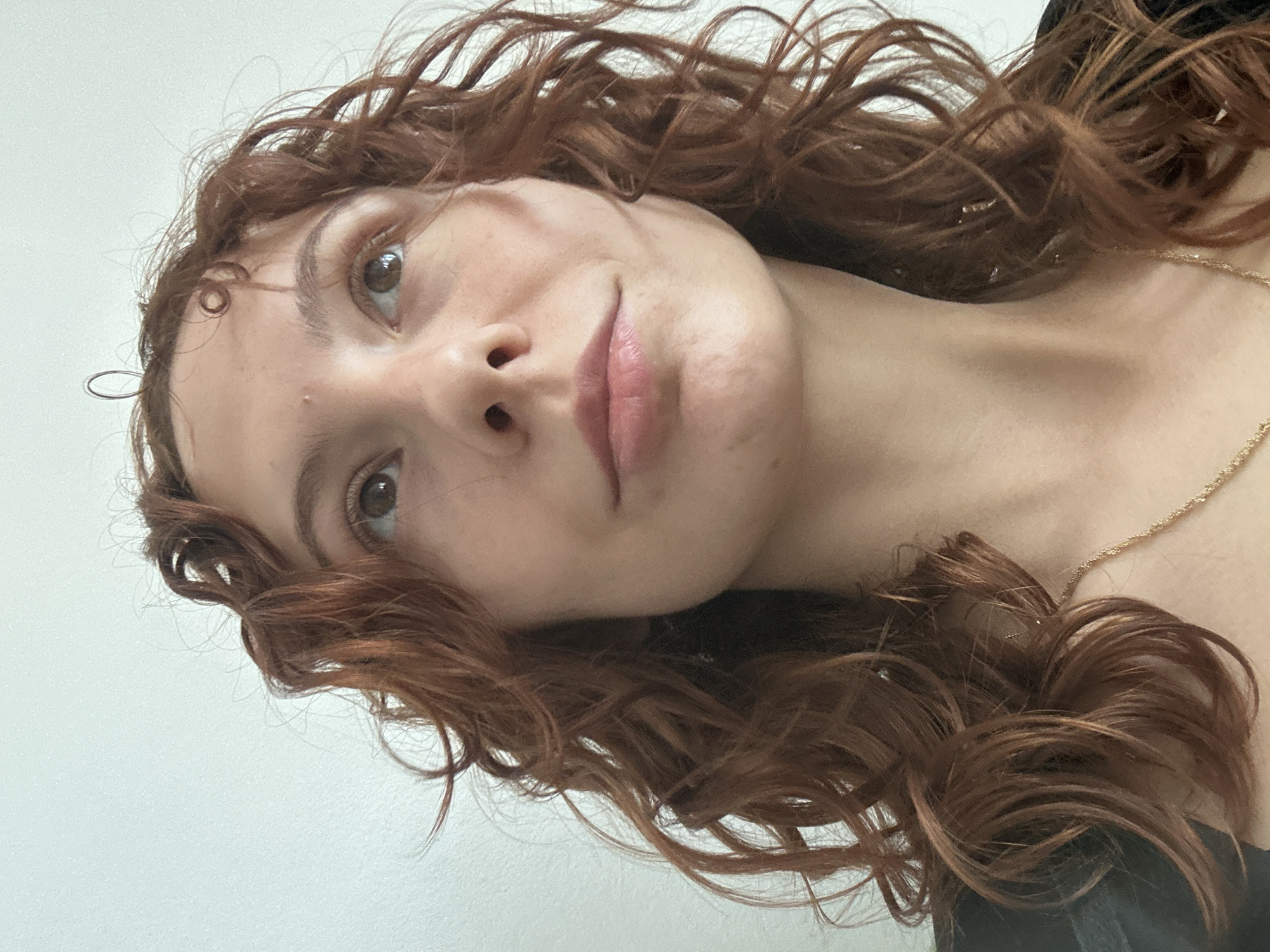 A close-up, tilted shot of a woman with brown curly hair and light skin. She is wearing a gold necklace. The background appears to be a plain, light-colored wall. No specific information indicates the location is in Novi Sad, Serbia.