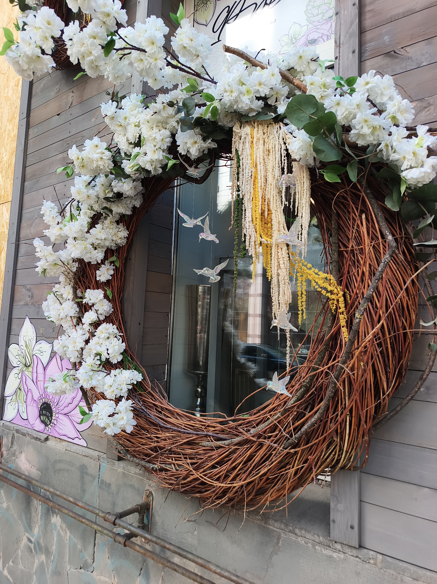 The image depicts an outdoor decorative display in what is likely a business storefront in Kyiv, Ukraine. A large, circular wreath made of interwoven brown branches dominates the scene. The wreath is adorned with white flowering branches, primarily concentrated on the left side and top of the circle. Hanging from the top of the wreath are strands of cream and yellow-colored decorative material, possibly tassels or fringe, interspersed with small, translucent bird-shaped ornaments. The wreath is mounted on a gray wooden wall. To the left of the wreath, part of a mural or sign featuring a large pink flower is visible on a concrete wall. The reflection visible in the window shows a blurred scene of the city. The image appears to have been taken during the day.