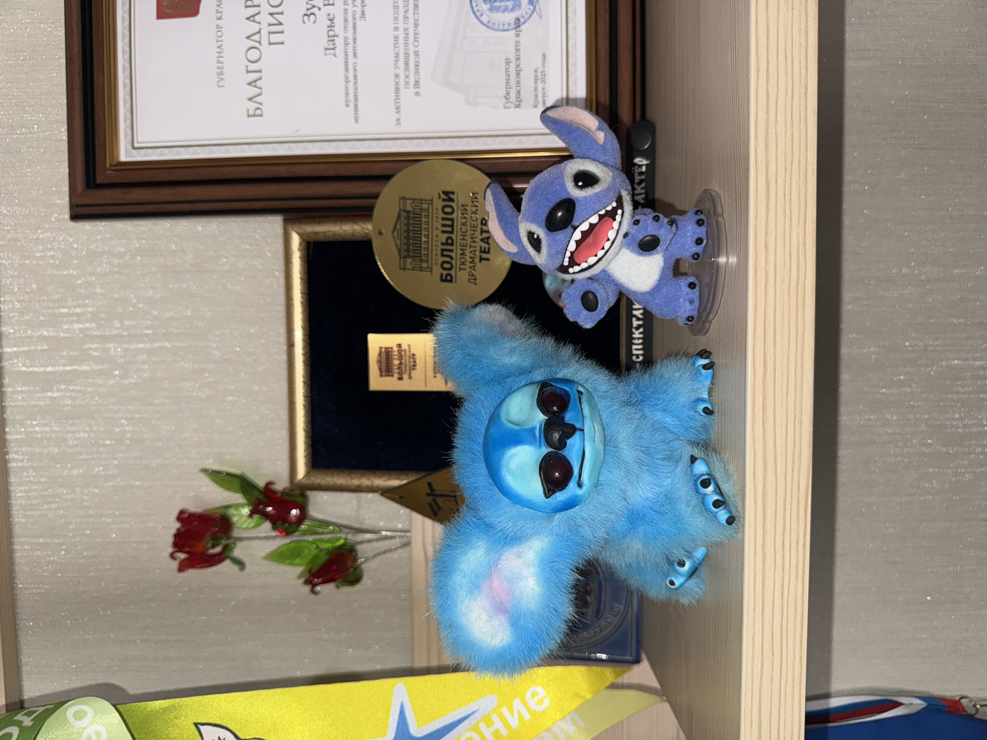 The image shows a shelf with several items on it. There are two plush figures of Stitch from the Disney movie "Lilo & Stitch." One Stitch is blue and furry, lying on its back, while the other is a blue figurine standing on a clear plastic base. Behind the Stitch figures are two framed documents. The top document is titled "БЛАГОДАР..." (BLAGODAR...) and mentions the Governor of the Krasnoyarsk Territory. It appears to be an award or certificate given to "Дарье В..." (Darye V...) for their role as a cultural organizer. The text mentions that the award is for active participation in events related to the Great Patriotic War. The second, gold-framed document contains text stating "БОЛЬШОЙ ТЮМЕНСКИЙ ДРАМАТИЧЕСКИЙ ТЕАТР" (BOLSHOI TYUMEN DRAMATIC THEATER). A small bouquet of artificial red roses is placed behind the blue, furry Stitch. The scene is likely located in Krasnoyarsk, Russia, as indicated by the document mentioning the Governor of the Krasnoyarsk Territory and the city itself.