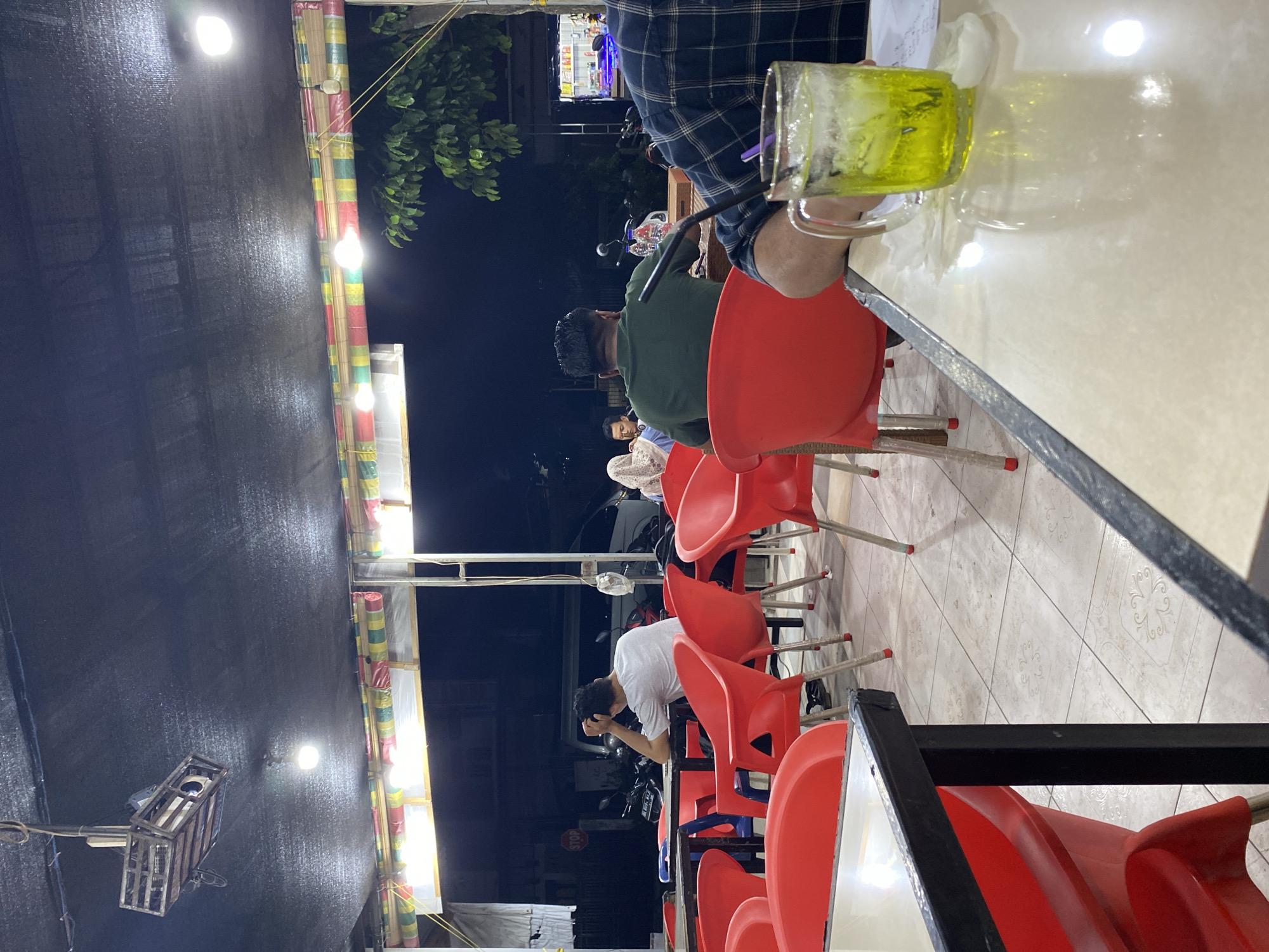 The image captures an evening scene at an outdoor dining area, likely a restaurant or cafe, in Lhokseumawe, Indonesia. The ambiance is casual and illuminated by artificial lights. In the foreground, a glass filled with a bright green beverage, possibly iced tea or a soda, sits on a table. The table surface is light-colored and reflective.
Several patrons are seated at tables. To the right, a person wearing a plaid shirt is partially visible, with their arm resting on the table near the drink. Further back, several individuals are seated at red plastic chairs. One man in a white t-shirt is seen from behind, with his hand on his head, appearing contemplative. Another person in a green t-shirt is also seated, facing away from the camera. More patrons, including a woman in a headscarf, are visible in the background, seated at tables.
The setting features an overhead structure covered with a dark fabric, from which lights are suspended, including a projector-like fixture. A decorative bamboo blind with colored stripes hangs along the edge of the covered area. Red plastic chairs are a prominent feature throughout the scene. The floor is tiled with a patterned design. In the background, motorcycles are parked, and a building with illuminated displays is visible, suggesting it is a commercial area. The overall impression is that of a relaxed, communal gathering space at night in the city.