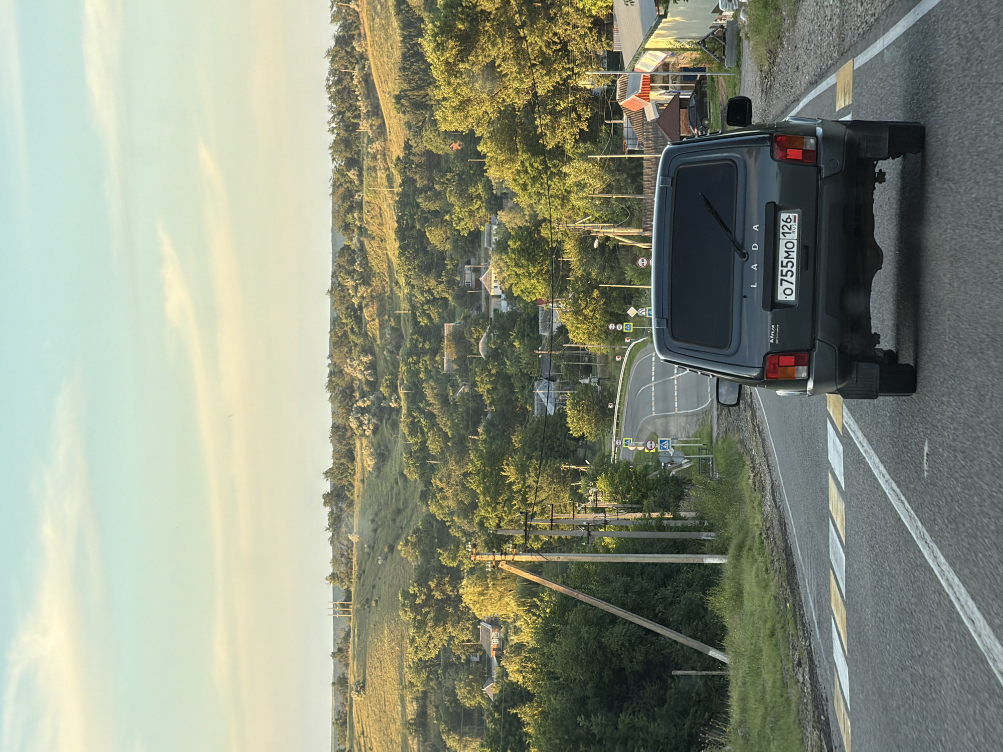 A dark gray Lada Niva car is driving on a paved road through a rural area. The car has a Russian license plate that reads "O755MO 126 RUS". The road has yellow and white pedestrian crossing markings. On the right side of the road, there are several buildings with red and white awnings. On the left side of the road, there are utility poles and trees. The background shows a rolling landscape with more trees and some houses, indicating a small village or town. The sky is a pale blue with some wispy clouds, and the light suggests it is late afternoon or early evening. The location is identified as Aleksandrovskoye, Russia, which is consistent with the rural scenery and the Russian license plate. There are no people visible in the immediate foreground or interacting with the car.