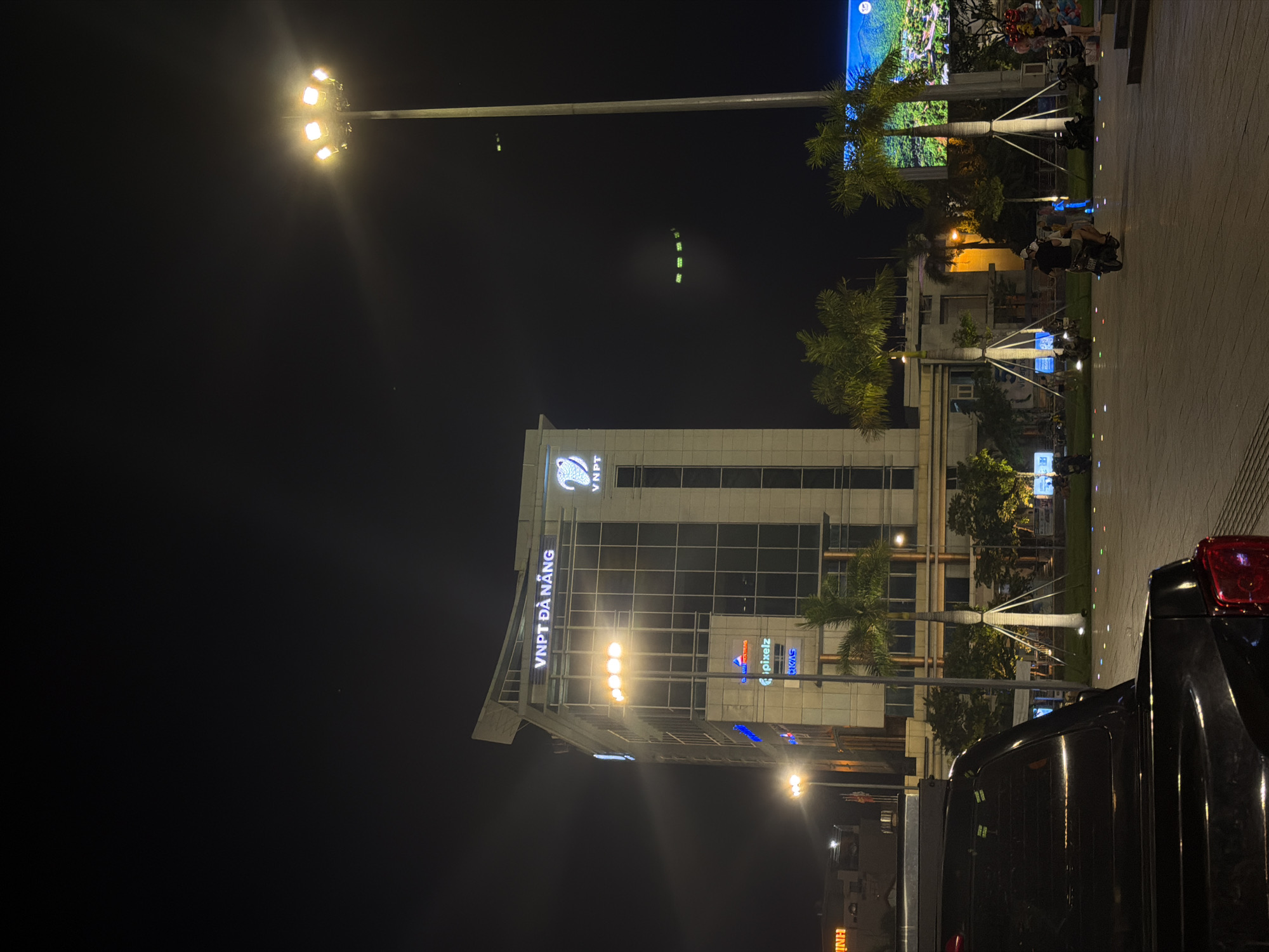 At night in Da Nang, Vietnam, a modern office building with illuminated signage "VNPT ĐÀ NẴNG" stands prominently. A large stadium floodlight illuminates the area from the right. In the foreground, the rear of a dark-colored pickup truck is visible. The scene is a public square with palm trees, streetlights, and a paved ground with colorful lights embedded in it. People on motorbikes are present, and a large digital billboard displaying a scenic landscape is visible in the background to the right. The overall atmosphere is urban and active, despite the late hour.