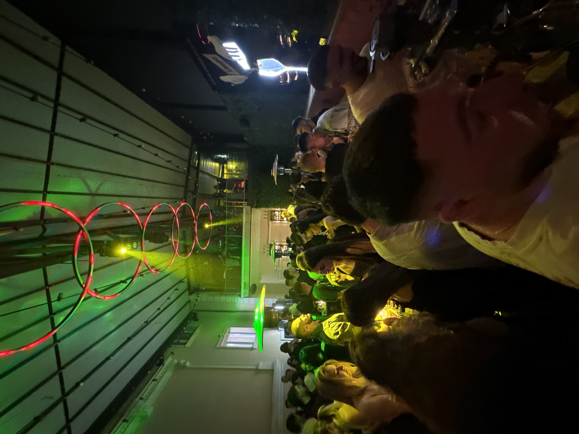 The image depicts a crowded outdoor event at night in Plovdiv, Bulgaria. A large group of people is gathered, facing toward a building illuminated with green light and red circular light fixtures. A sign with white lettering is visible above the crowd. The environment is dark, with the primary light sources emanating from the building and event lighting. People are closely packed, suggesting a social gathering or celebration.