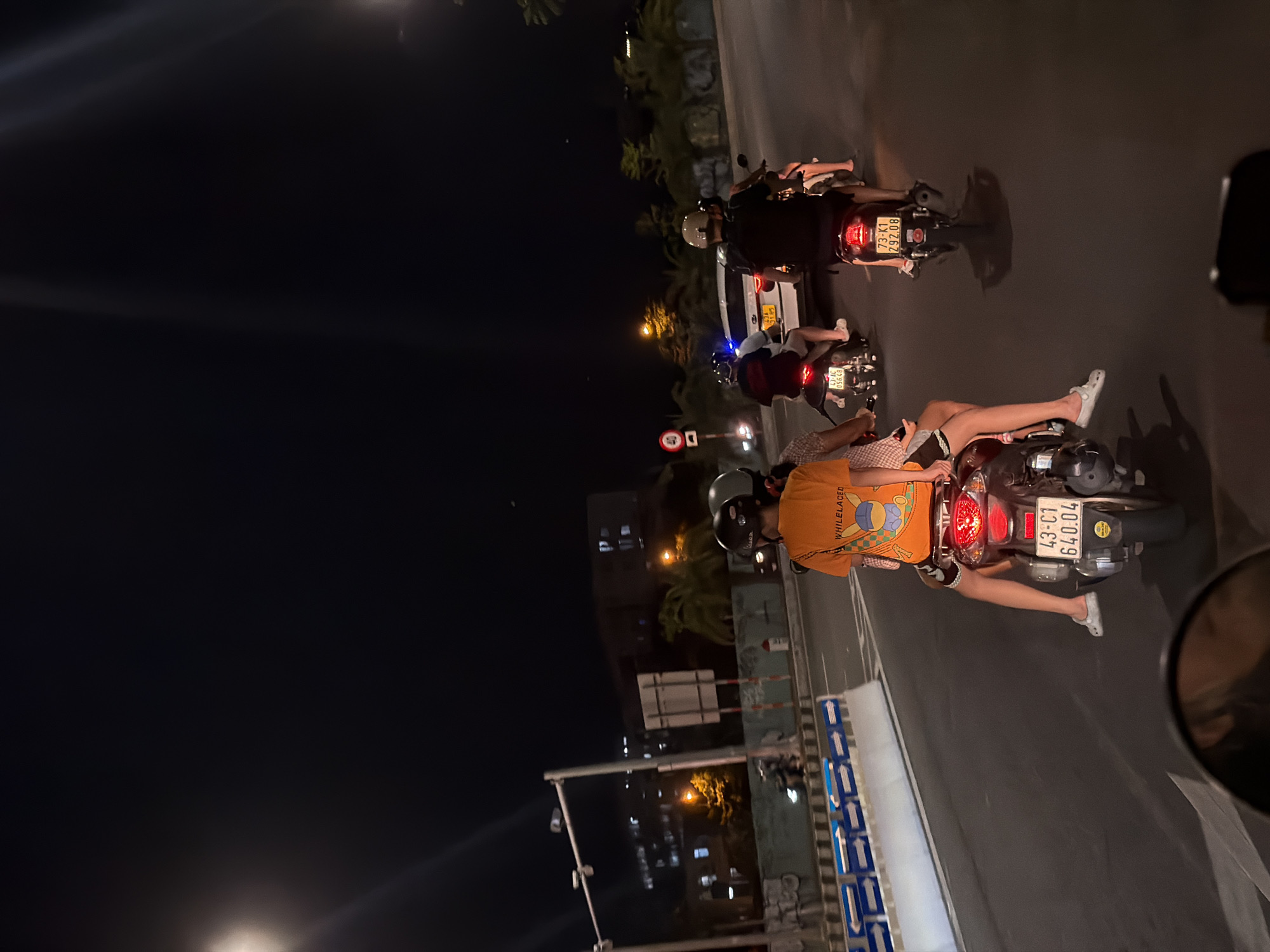 The image captures a nighttime scene on a city street in Đà Nẵng, Vietnam, likely a main road given the street lights and multi-lane traffic. The perspective is from a vehicle, possibly another motorbike, looking forward. In the foreground, a motorbike with two occupants is prominently featured. The rear passenger is a person wearing an orange t-shirt with graphics and dark shorts, their legs visible as they sit astride. The driver of this motorbike is wearing a helmet. Behind them, a second motorbike also carries two people, with the rear passenger wearing a dark shirt and the driver sporting a helmet. Further down the road, a third motorbike with a single rider wearing a helmet is visible. To the right, a white car is traveling in the same direction. The environment includes streetlights casting beams across the dark asphalt road, traffic barriers with blue and white directional markings, and buildings illuminated in the background. Palm trees are also present, suggesting a tropical climate. Traffic signs are visible, including a circular sign with a red border and a number 40 inside. The overall atmosphere is that of urban nightlife and transportation.