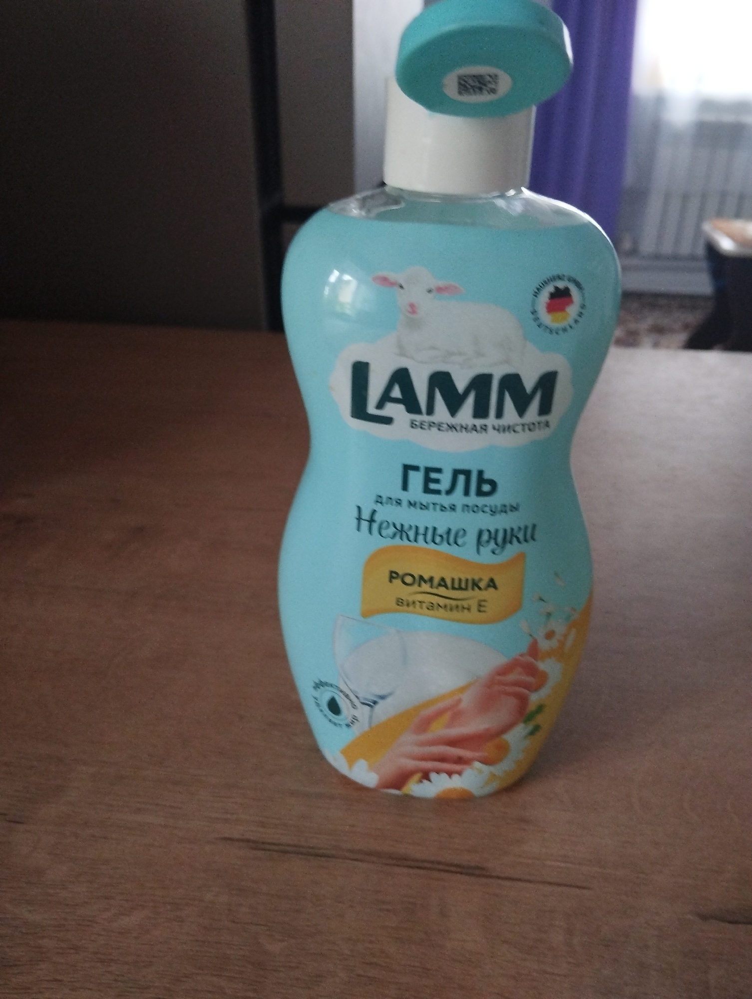 A close-up photograph displays a blue bottle of LAMM dishwashing gel standing on a brown wooden surface. The bottle features an image of a lamb at the top, followed by the brand name "LAMM" in bold letters. Below that, text in Russian indicates "gentle cleaning." Further down, the bottle describes the product as a "gel for washing dishes, gentle hands," and includes an image of chamomile with vitamin E. The cap of the bottle is a light blue color. The background is blurry but shows a dark brown cabinet and a window with curtains. The photograph appears to be taken indoors. There is no information in the image about the location in the city of Krasnogorskoye, Russia.