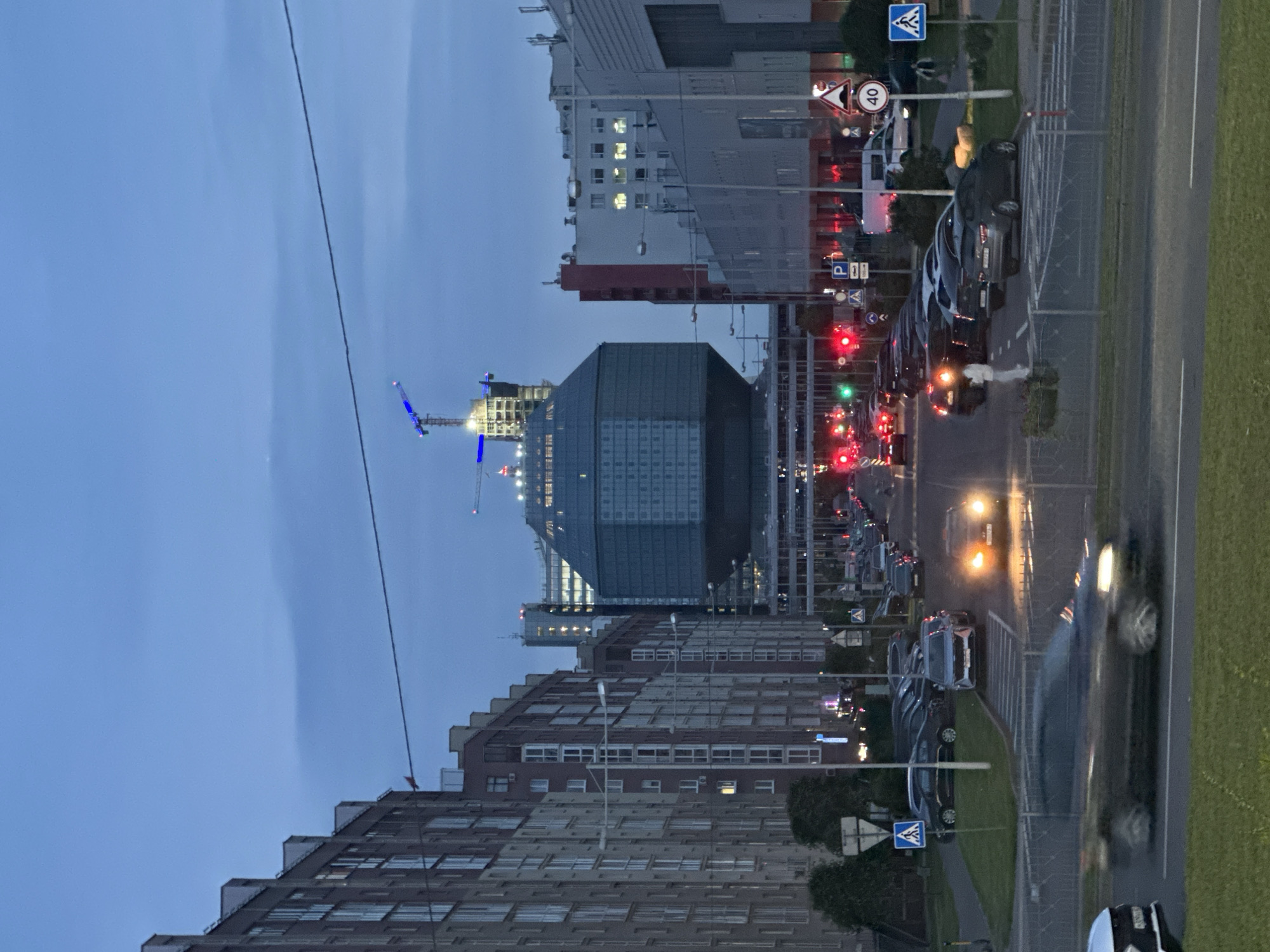 This is a vertical, medium-shot, dusk time photograph taken in Minsk, Belarus. The scene depicts a street with multi-story buildings on both sides. In the center of the frame, a large, modern building with a distinctive geometric, diamond-like shape, is under construction, topped with cranes. Cars are driving and parked on the street, and there are traffic lights and road signs visible. The sky is a deep blue, indicating twilight. The buildings on the left are residential, with many windows and balconies, while the buildings on the right appear to be commercial or office spaces. There is a pedestrian crossing with blurry figures of people crossing. The overall atmosphere is urban and active, despite the dim lighting.
