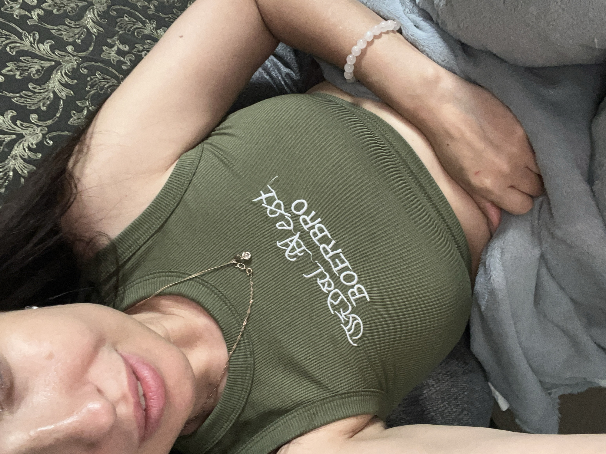 This is a close-up, overhead shot of a person lying down. The person is wearing a green ribbed tank top with white text on it that reads "deal with boerbo". They are also wearing a gold necklace with a small charm and a light pink beaded bracelet. The person's skin is fair, and they have dark hair. Their eyes are partially closed, and their lips are slightly parted, with a soft expression.
The person is lying on a patterned fabric, possibly a couch or bed, with a gray blanket draped over their body. The lighting suggests an indoor setting, likely during the day or with artificial lighting. The overall mood is relaxed and intimate. There are no obvious cues about the time of day, weather, or specific location.