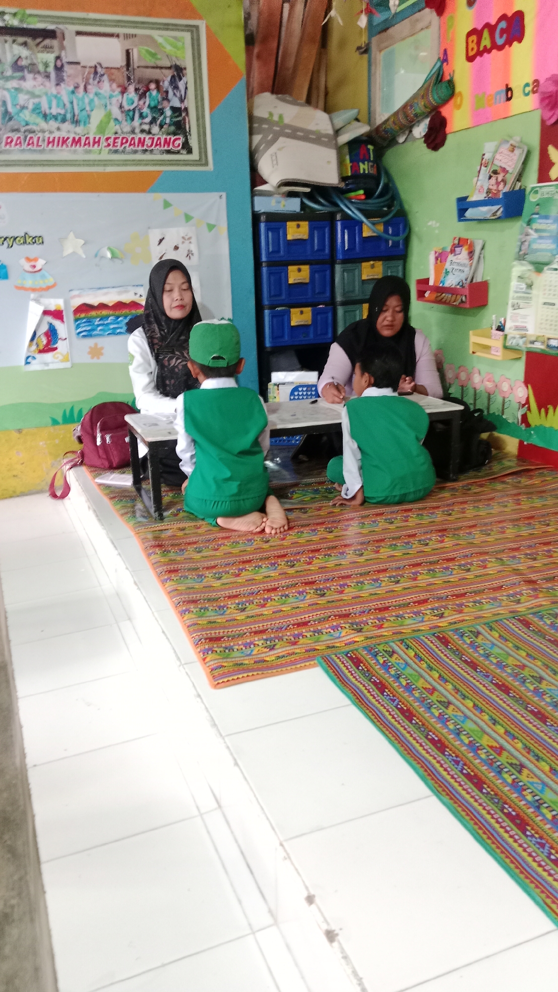 In a classroom in Karangharjo, Indonesia, two women wearing hijabs are seated with two young boys. The boys, wearing green shirts and hats, are kneeling on colorful rugs. One woman appears to be a teacher, leaning over a table and gesturing with her hand, possibly interacting with one of the boys. The other woman is seated behind the table, facing the boys. The room is decorated with children's artwork and educational materials, including a poster with the text "RA' AL HIKMAH SEPANJANG." Storage units filled with drawers are visible in the background, along with shelves holding books and other items. The floor is tiled, and the overall atmosphere suggests a learning environment for young children.