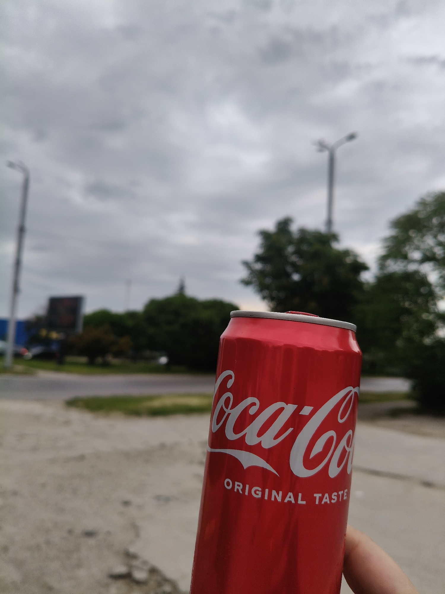 A close-up shot of a red and white Coca-Cola can with "ORIGINAL TASTE" written on it, slightly blurred background showing a street with a green median, buildings, trees, and overcast sky. Streetlights are visible in the background. The image was likely taken in an urban area, possibly Plovdiv, Bulgaria. The presence of street lights, buildings, and road infrastructure suggests a developed area within the city.