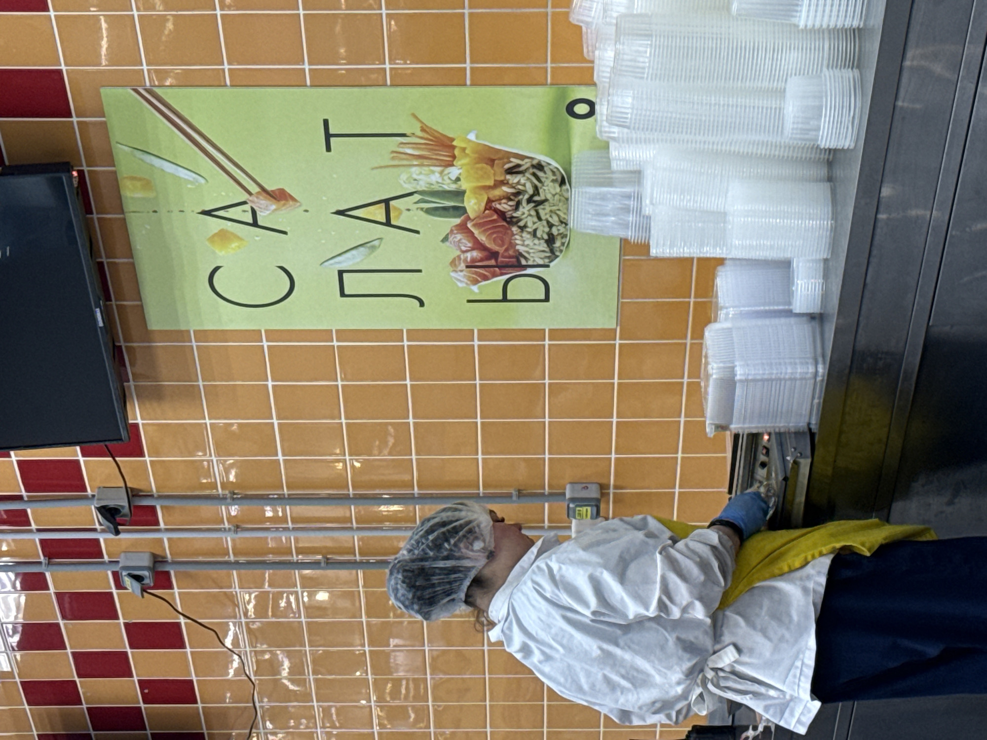 A view inside a food preparation area, possibly in Izhevsk, Russia. A worker wearing a hairnet, white coat, blue gloves, and dark trousers is standing in front of a stainless steel counter. To the worker's left is a poster showing a food bowl and the Russian word "Салат" (salad) with an image of chopsticks reaching for the ingredients. Next to the poster, atop the counter, is a stacked collection of transparent plastic food containers. The background features a wall with an orange and red tiled pattern. Electrical conduits and outlets are also visible on the wall.