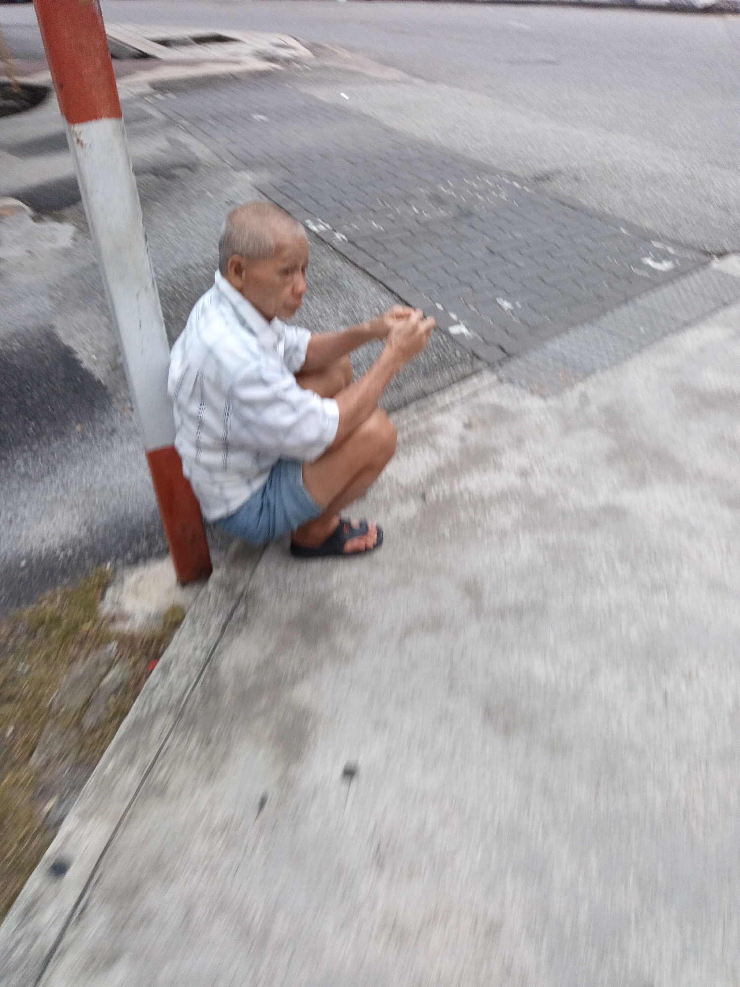 An elderly Asian man with sparse hair is squatting on a sidewalk next to a striped pole. He is wearing a white and blue striped short-sleeved shirt, blue denim shorts, and black sandals. His hands are positioned in front of him as if he is holding something or engaged in an activity. The background shows a gray paved road and a sidewalk with a textured surface. The scene is outdoors, and the lighting suggests it may be daytime. The blurriness of the image indicates movement.