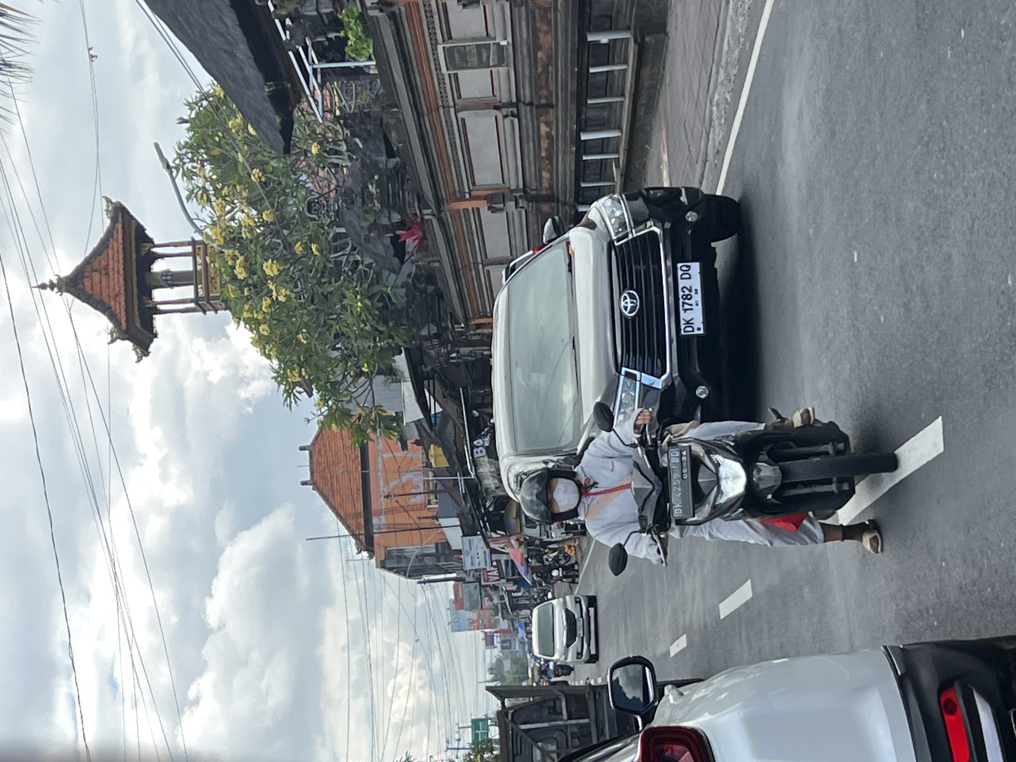 A person wearing a white outfit and a face mask rides a black motorcycle on a street in Abiansemal, Indonesia. The person is wearing a helmet and sandals. The motorcycle has a license plate that reads "DK 4259 FBD". The street is paved and has white lane markings. There are cars on the road, including a black SUV. Buildings line the street, with a traditional Balinese temple structure visible in the background. The sky is cloudy.