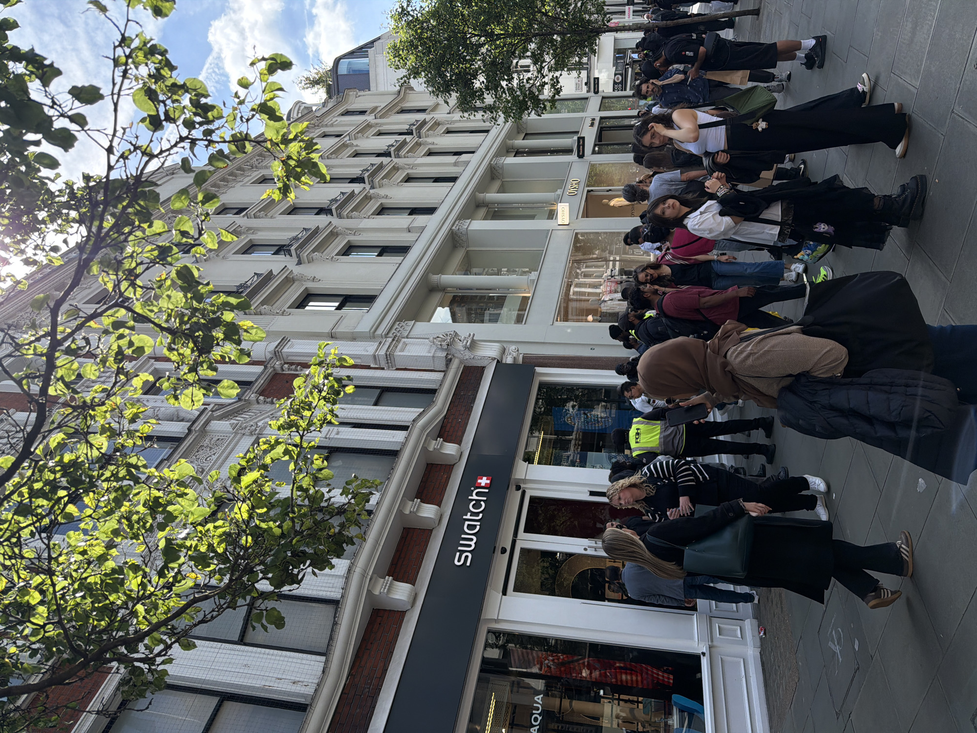 The image shows a daytime street scene in London, United Kingdom, with a queue of people outside a Swatch store. The store has a grey facade with the "Swatch" logo prominently displayed. People are standing along the sidewalk, forming a line that extends past the Swatch store towards other retail establishments, including one appearing to be a retail shop. The building is multi-story and made of white stone with architectural details. The sky is visible above the buildings, with scattered clouds. A tree with green leaves is in the foreground of the image. Pedestrians are dressed in casual attire. The sidewalk appears to be made of grey stone. The image suggests a crowd, likely waiting to enter the Swatch store.