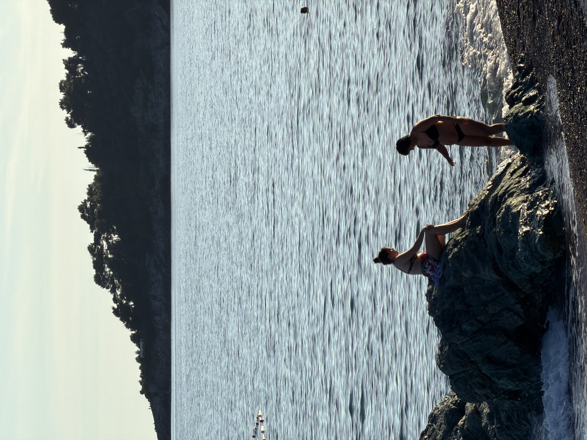 This media file captures a tranquil coastal scene in Rafailovići, Montenegro, focusing on two women by the water. The image appears to be taken on a clear day, with soft, natural lighting that suggests either early morning or late afternoon, as sunlight glints gently across the water.
In the foreground, a prominent dark, craggy rock formation juts into the calm sea from the right. One woman, clad in a dark bikini, stands on a lower section of these rocks, where small waves lightly break. She is leaning forward, looking down towards the water, possibly observing it or preparing to enter. Slightly higher and closer to the viewer on the same rock structure, another woman is seated comfortably. She wears a patterned swimsuit and has her hair tied up, adopting a relaxed posture with her legs drawn up, indicating a moment of repose or contemplation amidst the natural beauty.
The expansive body of water, likely the Adriatic Sea, dominates the midground, showcasing subtle ripples and reflections of light. In the distance on the water, several small, light-colored objects, resembling buoys or small vessels, are scattered, along with one larger dark object further out. The left side of the background reveals a lush, dark green hillside densely covered with trees, forming a natural boundary against a bright sky. A solitary white building, possibly a house or a small hotel, is visible on the distant forested slope.
The overall mood conveyed is one of peace and relaxation, highlighting a leisure activity in a picturesque natural setting. There is no visible text in the image.