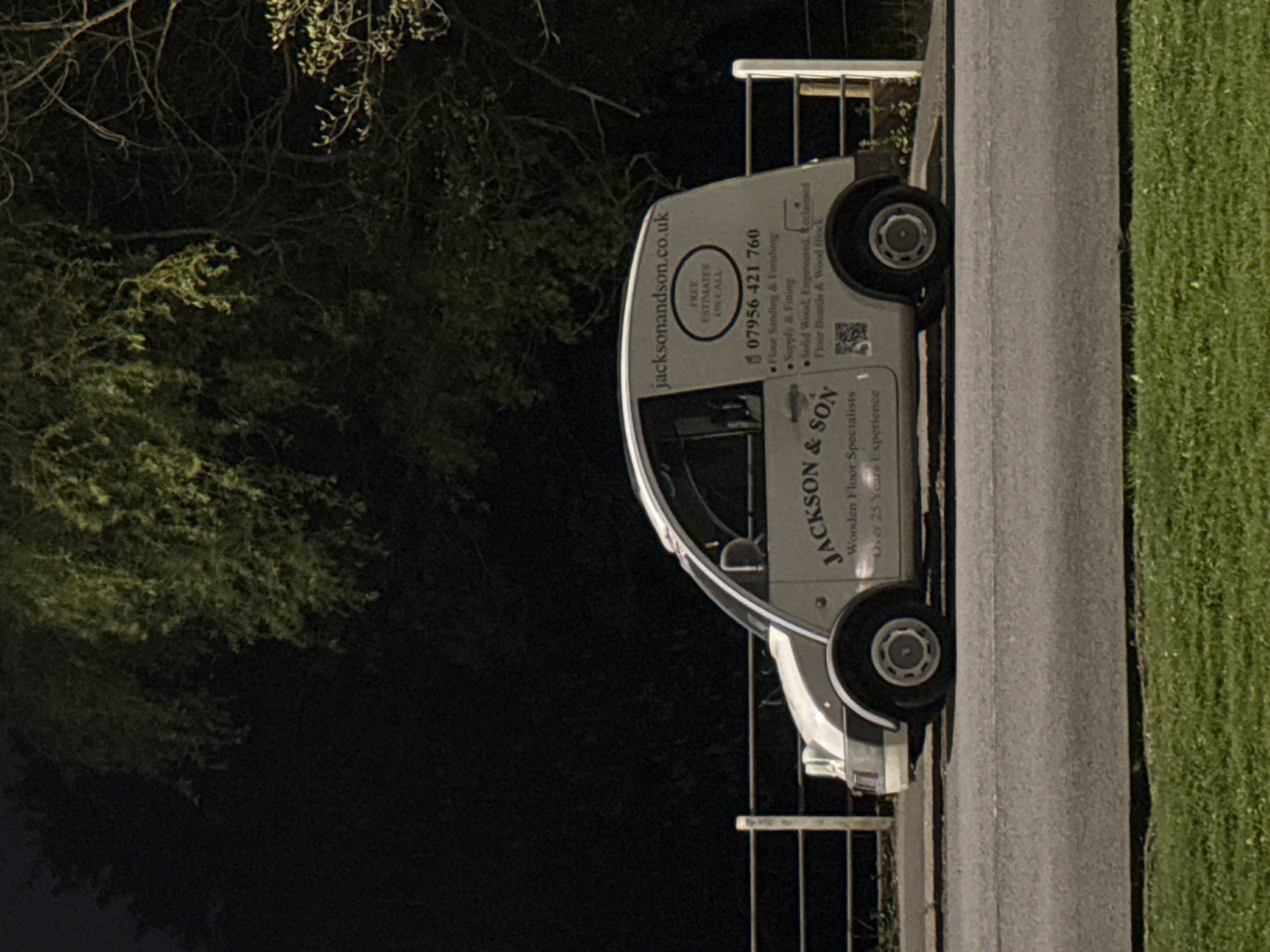The image, rotated 90 degrees clockwise, displays a white commercial van on an asphalt road or path, flanked by a strip of green grass on the right. To the left and above the van, dark, leafy trees and tangled branches are visible, suggesting an outdoor setting during low light conditions, such as dusk, night, or early morning.
The side of the van is clearly marked with business information for "JACKSON & SON," specializing as "Wooden Floor Specialists" with "Over 25 Years Experience." Contact details include the website "jacksonandson.co.uk" and a UK phone number, "07956 421 760." Services offered are "Floor Sanding & Finishing," "Supply & Fitting" of various floor types including "Solid Wood, Engineered, Reclaimed Floor Boards & Wood Block." A circular emblem advertises "FREE ESTIMATES ON-CALL." The UK-specific contact information is consistent with the provided context of London, United Kingdom. No individuals are present in the scene.