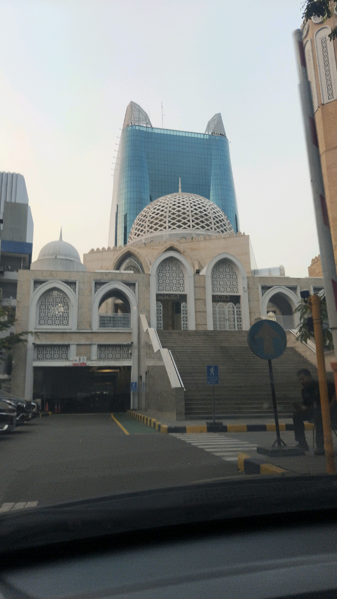 An exterior, low-angle, eye-level, medium shot shows the exterior of the IDX Mosque in Central Jakarta, Indonesia, with a modern blue skyscraper in the background. The mosque has a large, ornate dome and multiple arched windows with intricate latticework. A wide staircase leads up to the mosque entrance. In the foreground, a street with a pedestrian crossing and traffic barriers is visible. A person is standing near a yellow pole on the right side of the image. The sky is overcast and pale blue. The overall impression is one of contrast between traditional Islamic architecture and modern urban development.