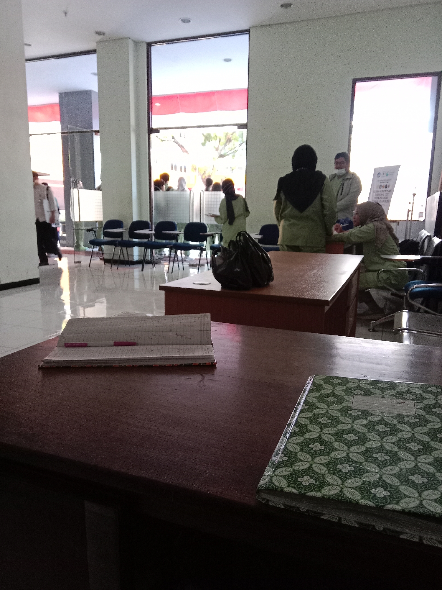 The image depicts an indoor scene, possibly an office or waiting area, with a bright, reflective tiled floor and light-colored walls. Large windows with red and white banners are visible in the background, suggesting a public building. In the foreground, two wooden tables are present. The closest table has an open notebook with a pink pen and a green patterned notebook on it. The second table, further back, holds a dark plastic bag. Several people are in the middle ground, some wearing light green uniforms and headscarves, indicating they might be employees or attendees. Some are standing, while others appear to be seated in blue chairs arranged around smaller tables. A man in a white shirt and dark pants is seen walking away from the camera on the left side of the frame. The overall ambiance suggests a functional, somewhat sterile environment. The presence of a red and white banner, common in Indonesian public spaces, and the attire of some individuals hint at the location being in Indonesia, potentially within the city of Malang.