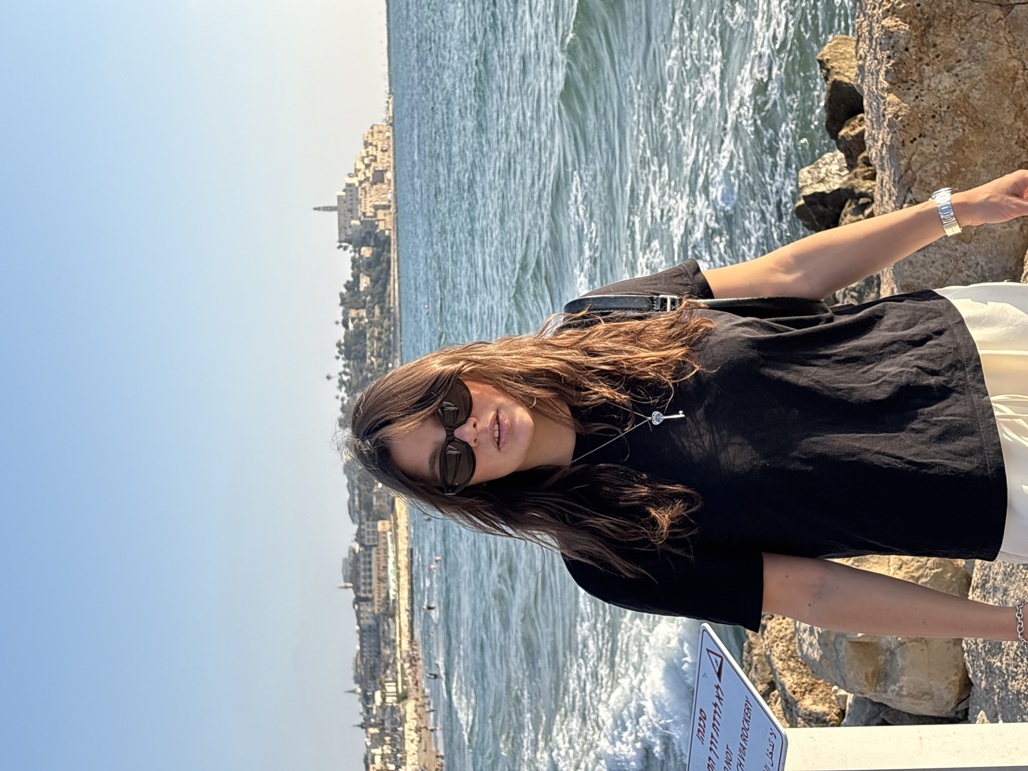 A woman with long brown hair and sunglasses stands on a rocky shore with the Mediterranean Sea in the background. She is wearing a black t-shirt, a silver necklace with a key pendant, and a silver watch. The sea is choppy with waves breaking against the shore. In the distance, a city skyline with buildings and a tower is visible. A sign in Hebrew and English is partially visible in the foreground to the left. The scene appears to be Jaffa, an ancient port city that is part of the larger Tel Aviv-Yafo municipality in Israel, known for its historic architecture and picturesque coastline.
