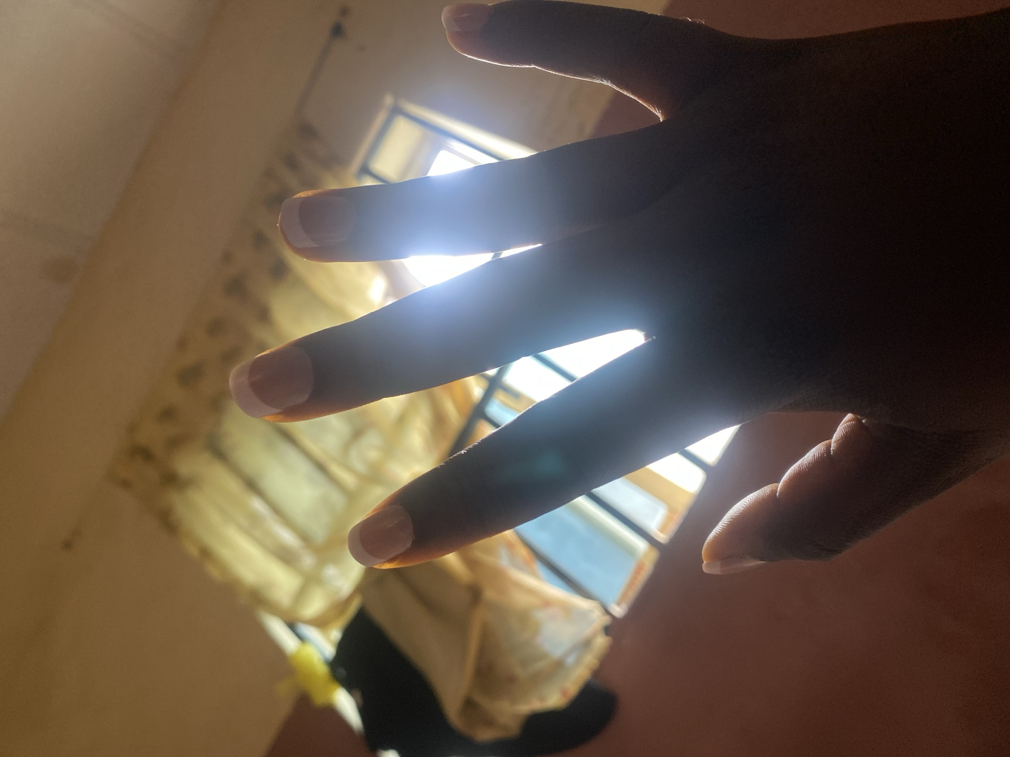 The image shows a close-up of a person's hand, with their fingers extended upwards towards a bright light source, likely sunlight streaming through a window. The hand is silhouetted against the light, obscuring most of the details of the fingers and palm. The fingernails appear to have a French manicure. In the background, a blurry window with panes is visible, and behind that, some fabric, possibly curtains, is also out of focus. The overall impression is a dimly lit interior with strong backlighting. The image does not contain any information that allows for the determination of its location in Jauro Yinu, Nigeria.