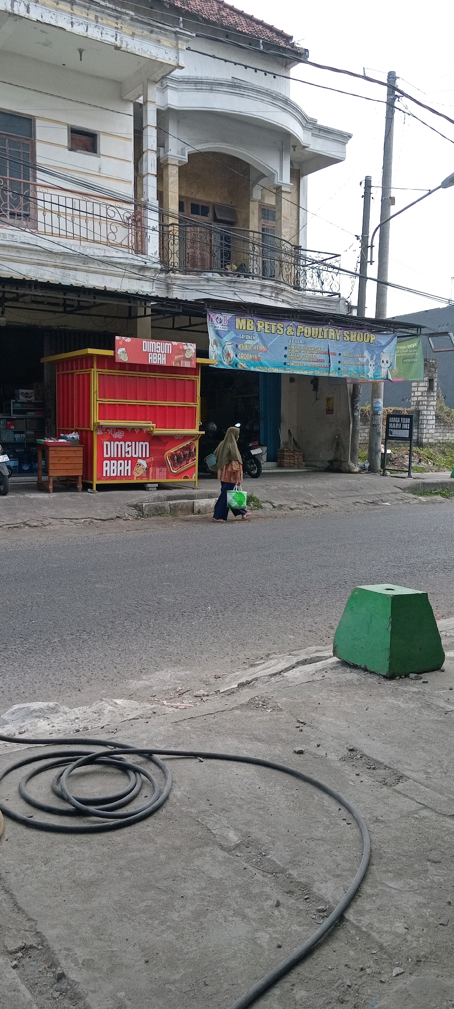 The image depicts a street scene in Babat, Indonesia. In the foreground, a coiled black hose lies on a concrete surface. Adjacent to the street, a red food stall with "DIMSUM ABAH" signage is visible. A woman wearing a hijab and carrying a green bag walks past the stall. Behind the stall, a banner for "MB PETS & POULTRY SHOOP" is displayed on a building. The building itself appears to be a multi-story structure with balconies and windows. The environment is urban, with a paved road, sidewalk, and power lines overhead.