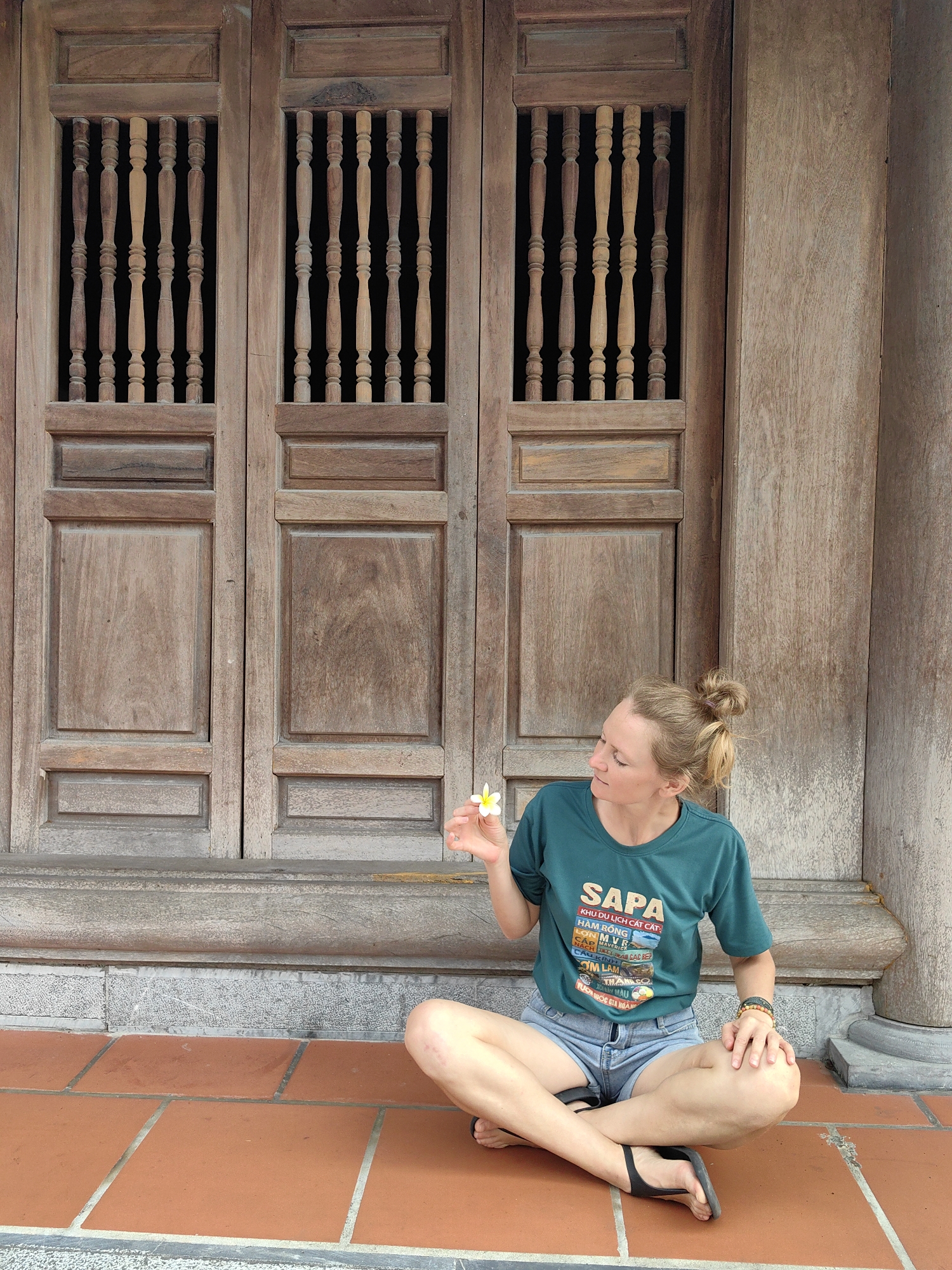 A woman is seated cross-legged on a reddish-brown tiled floor, positioned against a traditional dark wooden facade in Hong Gai Ward, Vietnam. She holds a small white and yellow flower in her right hand and is looking down at it with a calm, contemplative expression. Her posture suggests a moment of rest or quiet observation.
The background features a large, weathered wooden structure, likely an intricately crafted door or wall of a building. It consists of multiple panels, with the upper sections featuring rectangular openings filled with vertical, turned wooden balusters that allow a view into a dark interior. A sturdy wooden pillar is visible on the far right, suggesting part of a larger architectural structure. The floor is composed of large, square, terracotta-colored tiles with contrasting grey grout lines.
The woman has light-colored hair pulled up into a messy bun. She is wearing a dark teal short-sleeved t-shirt and light blue denim shorts, along with black flip-flops. A beaded bracelet adorns her left wrist. The t-shirt features prominent white and colored text in Vietnamese, which includes "SAPA KHU DU LỊCH CÁT CẤT HÀM RỒNG," followed by lines like "LON MVR," "NÁCH TITAN CAC BE," "MAVE," "CÂU KÍNH," "OM LAM," "THAN CO," and "TUON QUOC GIA HOÀNG." The scene is brightly and evenly lit, indicating daytime conditions, contributing to a serene atmosphere.
