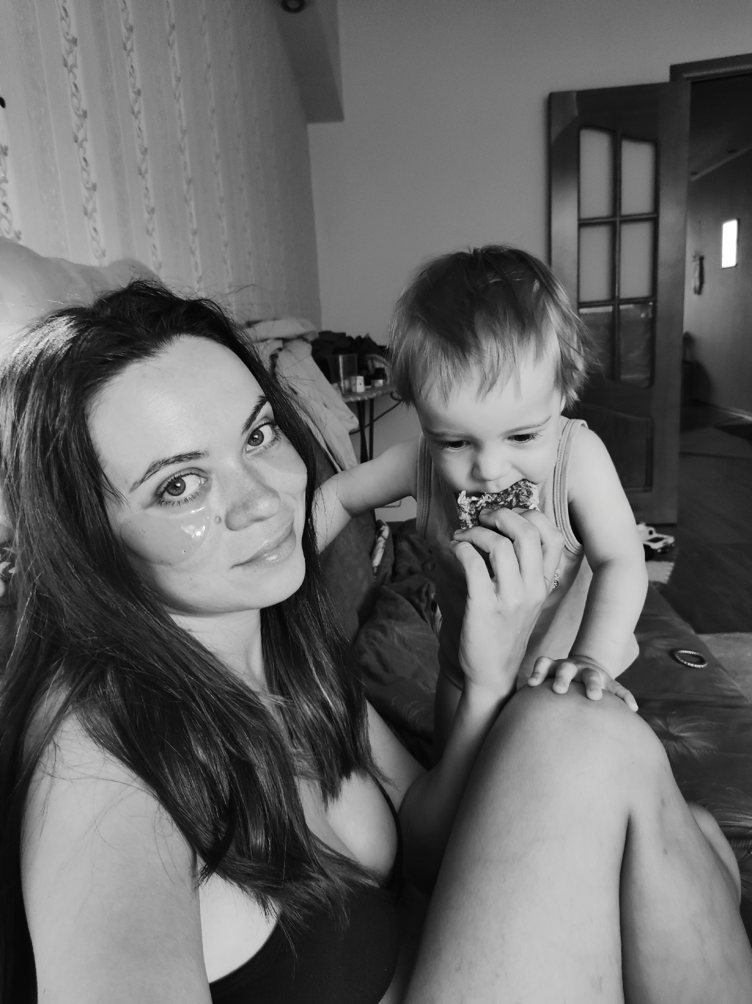 A black and white medium shot shows a woman and a young child inside an apartment in Chaikovsky, Russia. The woman, with long hair, is wearing eye masks under her eyes and is looking at the camera. She is sitting on a surface that appears to be covered in a dark material. The child, who appears to be no more than two years old, is standing near the woman and eating something from her hand. A partially open door is in the background. Additional furniture is visible. The scene is lit with natural light.