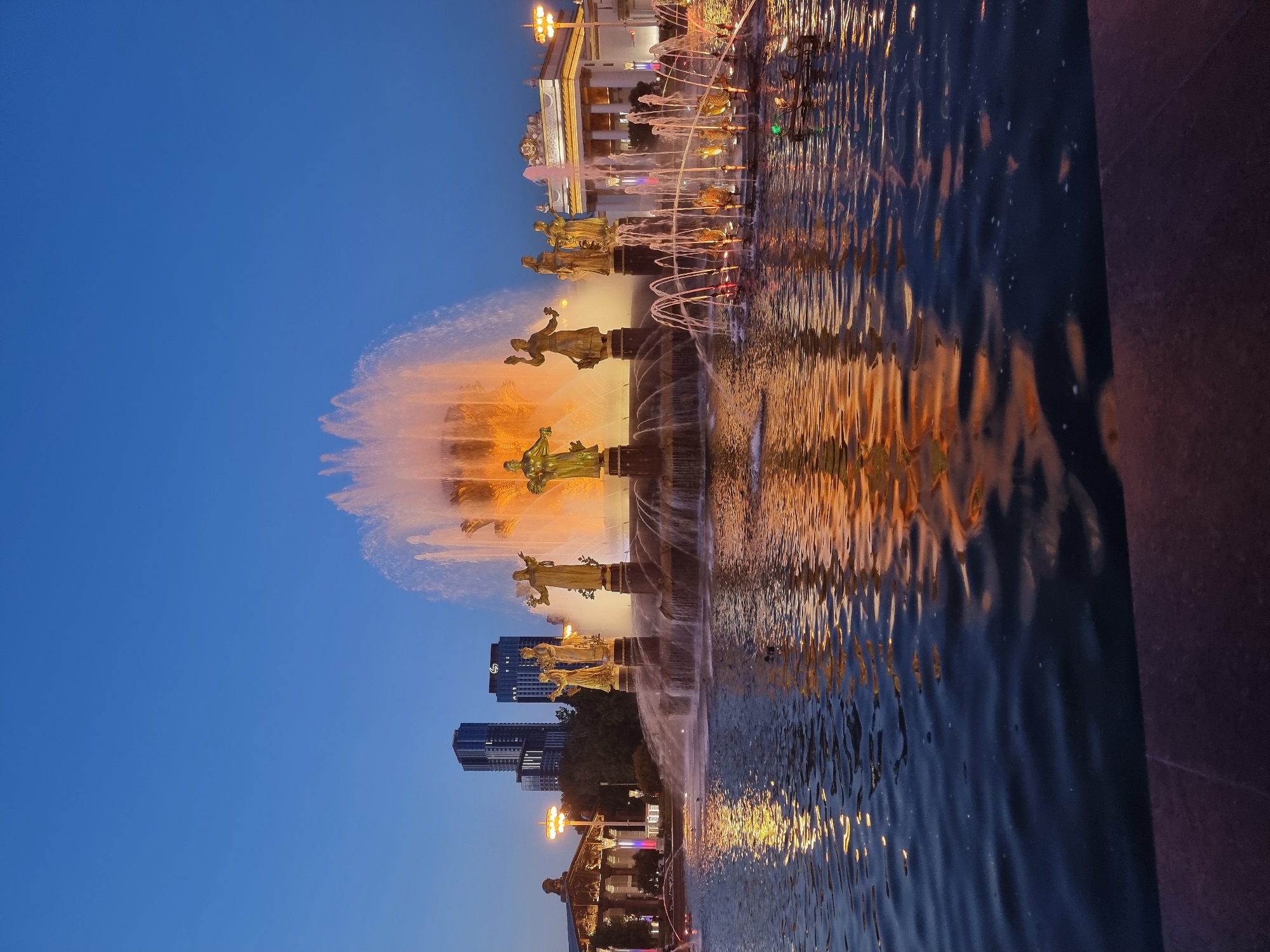 The image captures the Friendship of the Peoples Fountain in Moscow, Russia, at dusk. The iconic golden statues of allegorical figures representing Soviet republics are illuminated, standing atop the fountain's tiered structure. Water cascades down the levels and erupts in a central spray, catching the warm orange and yellow lights from the fountain's illumination. The water's surface reflects the lights and the golden statues, creating a shimmering effect. In the background, modern skyscrapers rise above a classic, ornate pavilion, also illuminated with lights. The sky is a deep, clear blue, indicating the transition from day to night. No people are visible in the frame. The overall scene conveys a sense of grandeur and artistic splendor.