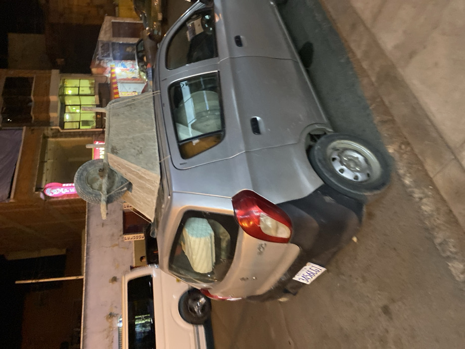 The image depicts an urban street scene at night in La Paz, Bolivia. A silver Suzuki Alto hatchback, license plate 3456LGT (Bolivia), is observed from its rear-right quarter. An inverted, concrete-stained wheelbarrow is secured to the vehicle's roof, with its single wheel positioned towards the front. Multiple rolls of light-colored material are visible inside the car through the rear passenger and tailgate windows. The vehicle's rear bumper is composed of black, unpainted plastic. In the background, a white van is partially visible to the left, and additional vehicles and multi-story buildings with illuminated windows are present further down the street. The scene is illuminated by artificial light sources. No individuals are clearly discernible.